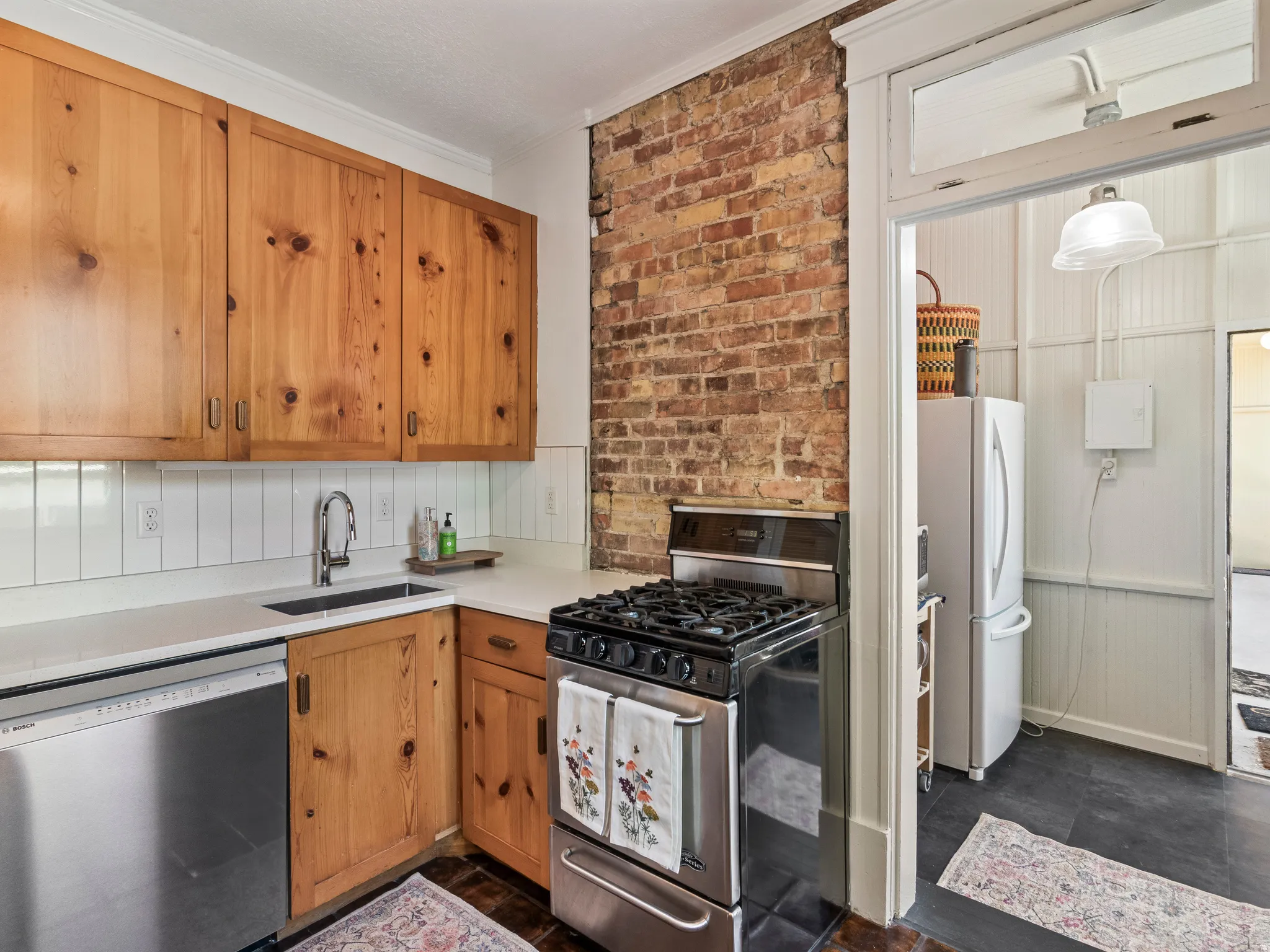 Kitchen featuring natural wood cabinetry, exposed brick accent wall, white subway tile backsplash, stainless steel gas range, and a built-in dishwasher