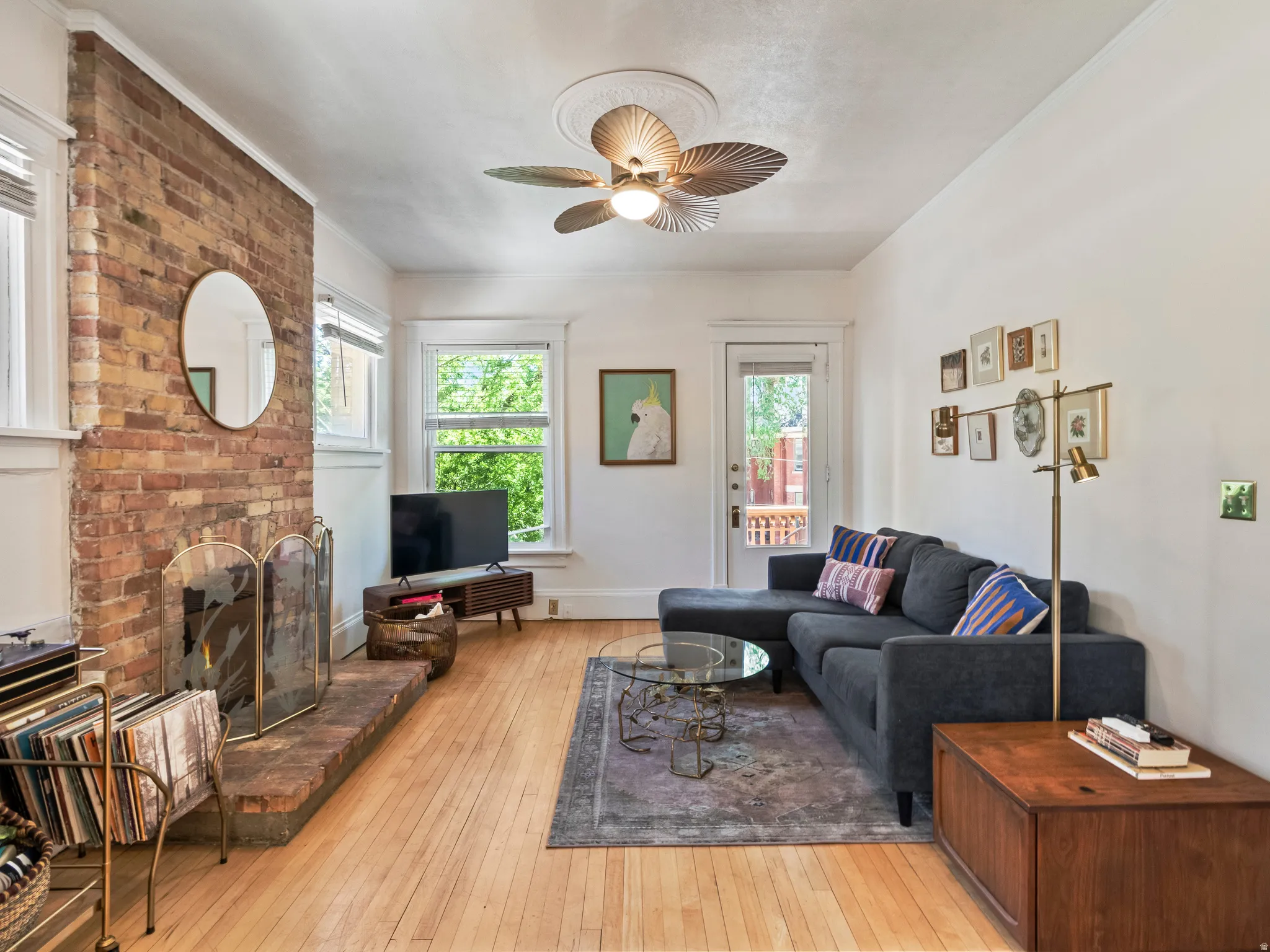 Exposed brick fireplace with hearth, wood-finish flooring, white trim windows, ceiling fan with light fixture, and a glass-paneled door