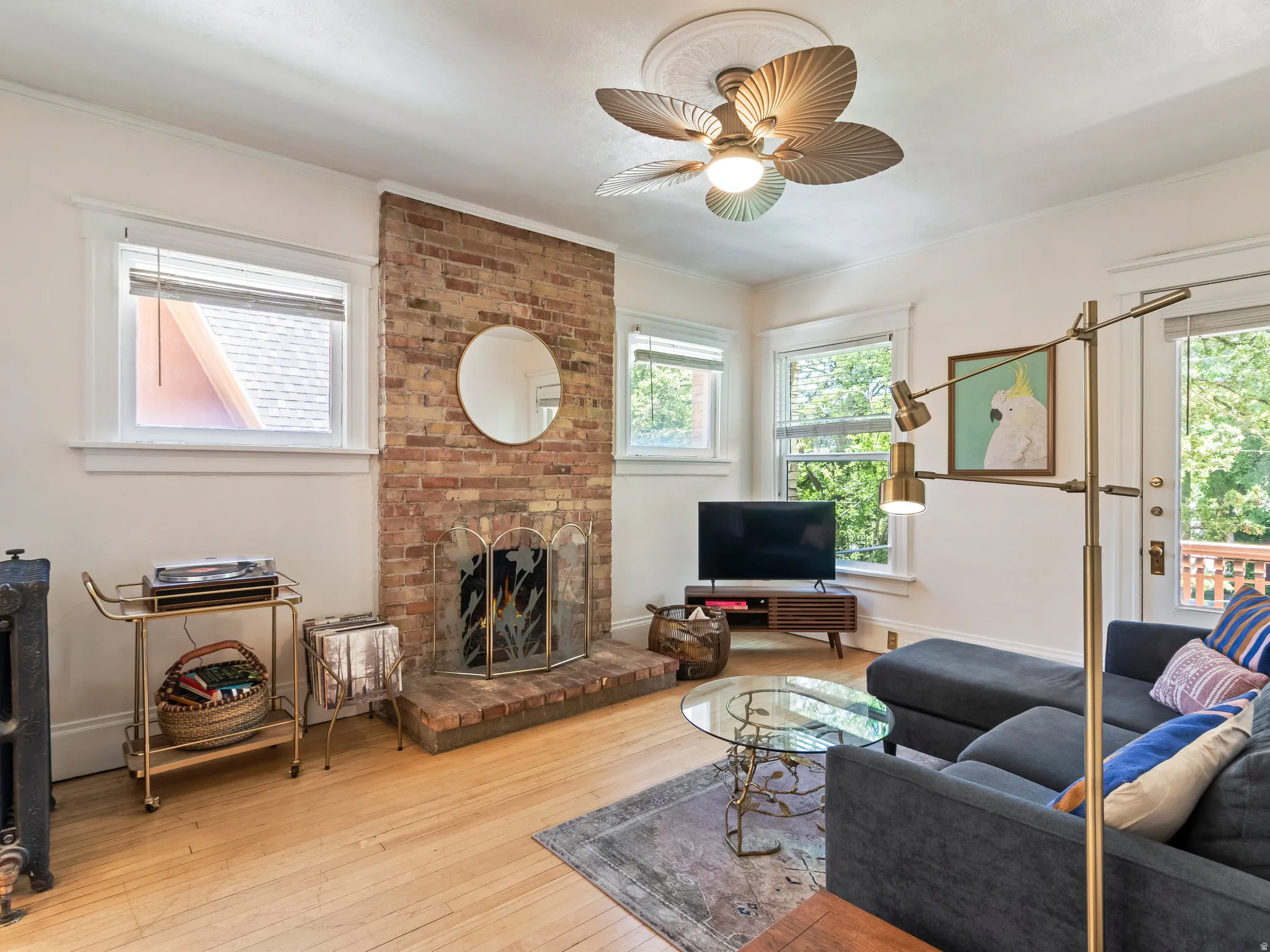 Living area featuring a brick fireplace with a metal screen, light wood-finish flooring, multiple windows, a ceiling fan with a medallion, and a white trim package