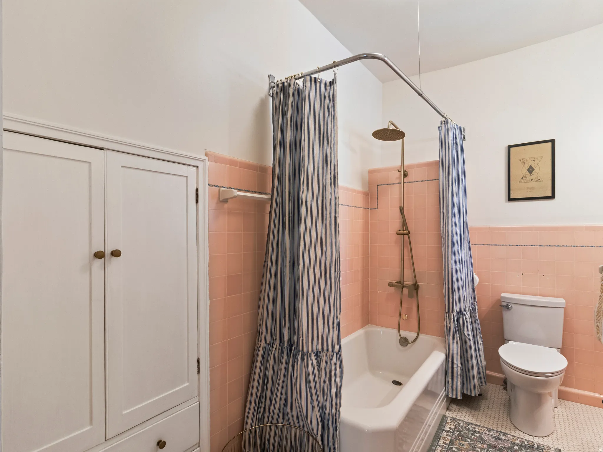 Bathroom featuring a clawfoot tub with an overhead shower fixture, pink tiled walls, a white toilet, and white built-in cabinetry with wood knobs