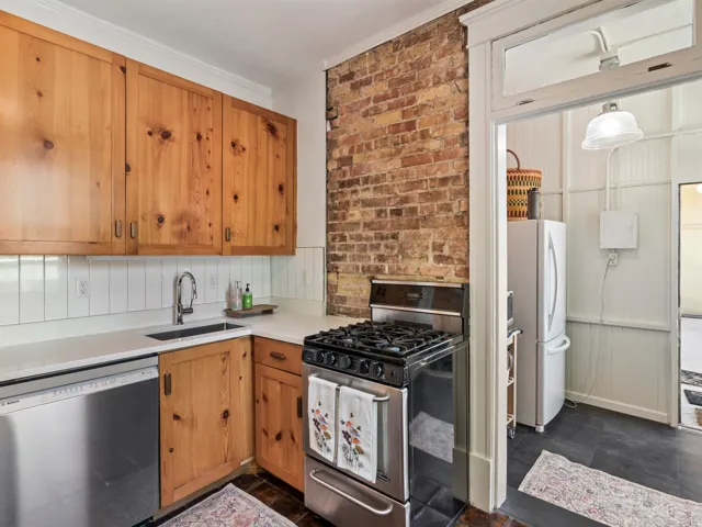 Kitchen featuring natural wood cabinetry, exposed brick accent wall, white subway tile backsplash, stainless steel gas range, and a built-in dishwasher