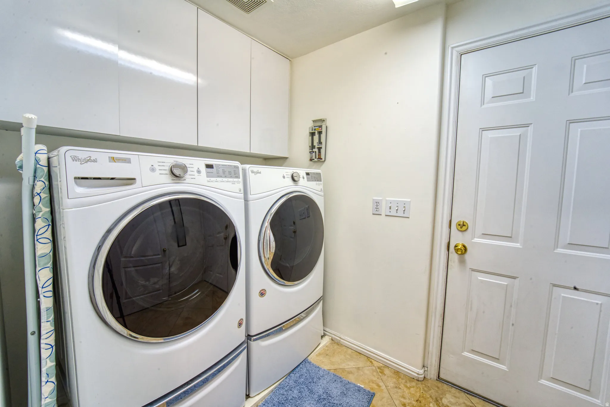 Dedicated laundry area featuring a white front-loading washer and dryer set, ample overhead white cabinetry for storage, a white paneled door with brass hardware, and tiled flooring