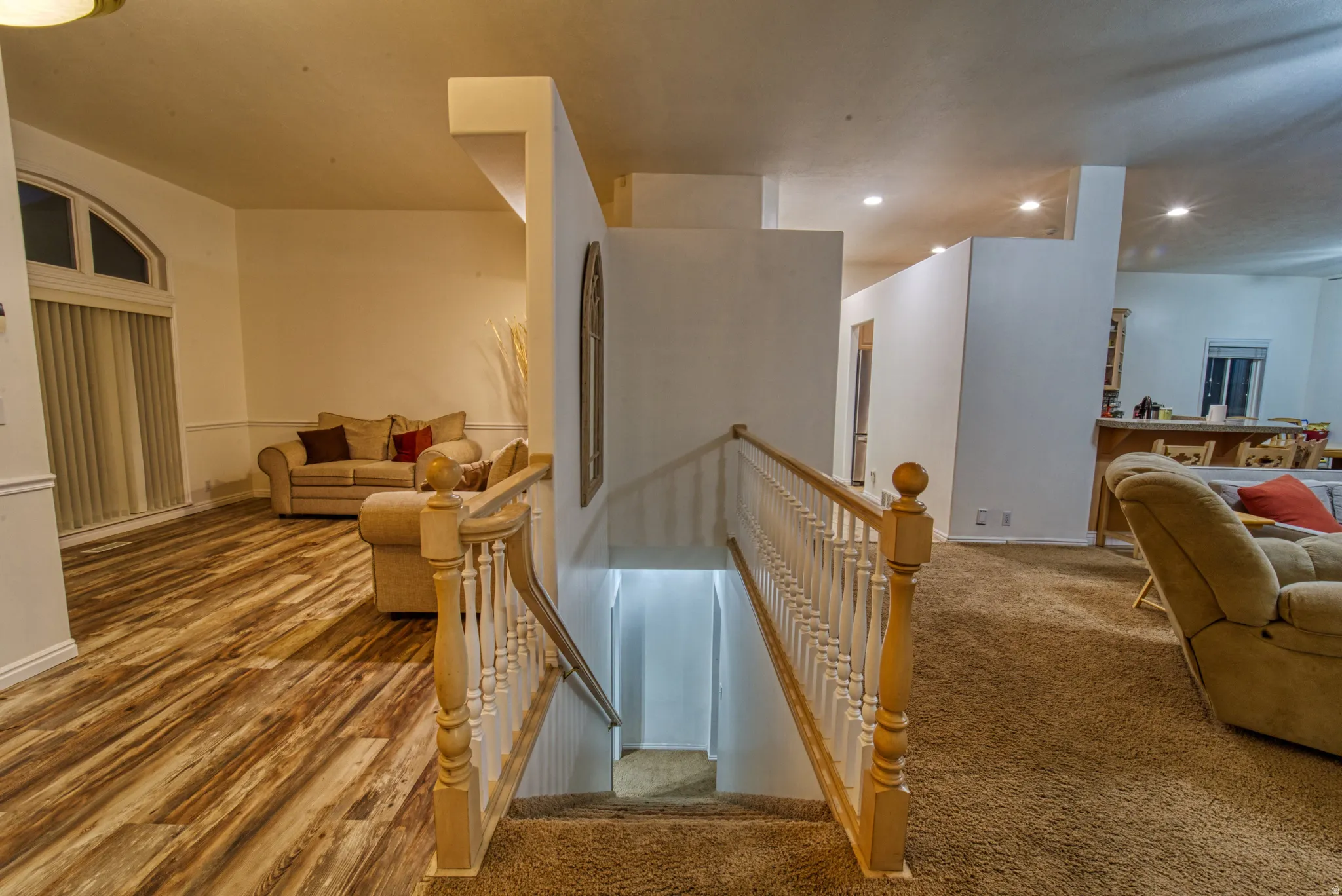 Split-level interior featuring wood-finish flooring, recessed lighting, white baseboards, a carpeted staircase with a wood handrail and white balusters, and a large arched window with vertical blinds