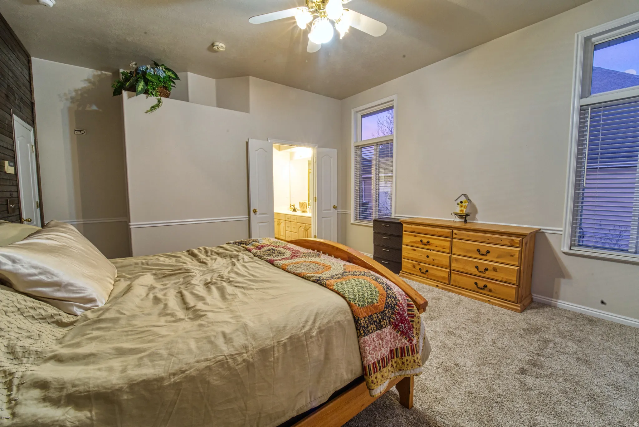 Carpeted bedroom featuring a textured accent wall, two windows with blinds, a ceiling fan with integrated lighting, and an open doorway revealing a bathroom with a dual vanity