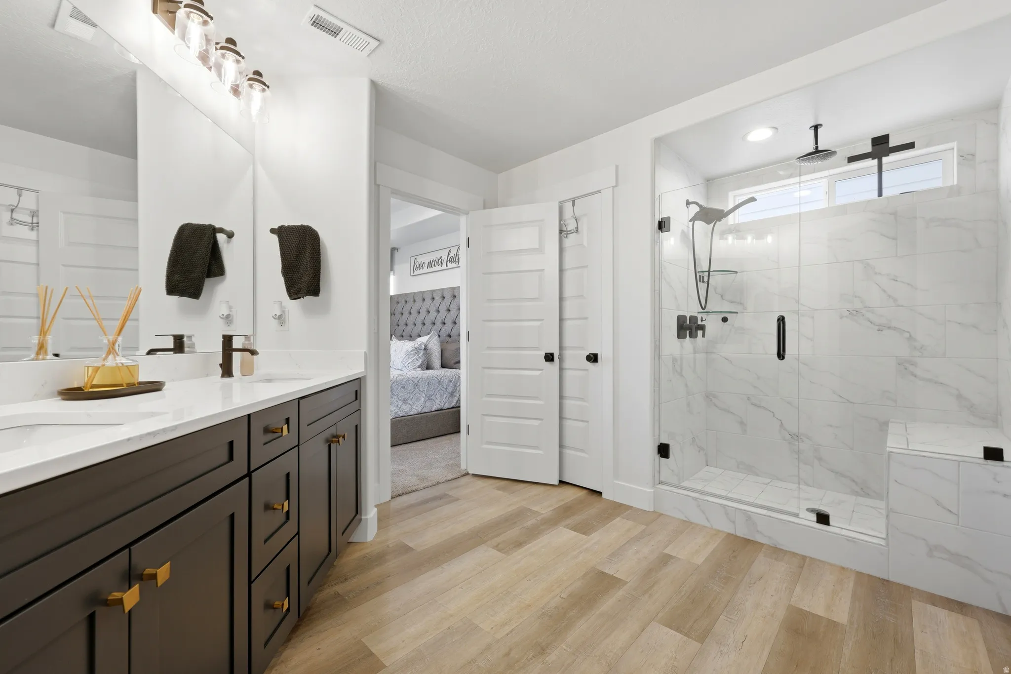 Spacious bathroom featuring wood-finish flooring, a dual vanity with dark cabinetry and white countertops, a large walk-in shower with glass enclosure and marble-look tile, and modern black fixtures