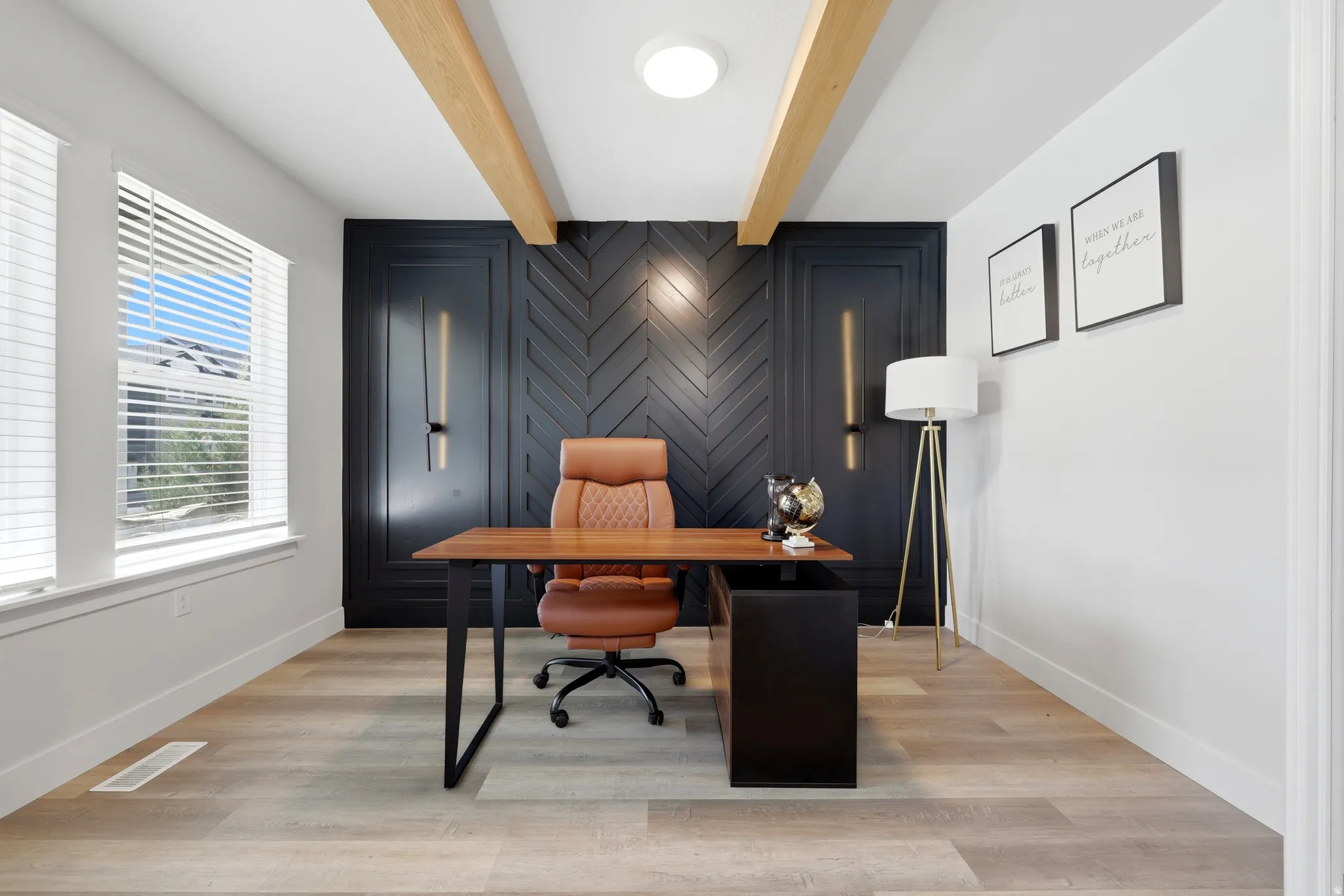 Office space featuring a dark accent wall with a herringbone pattern, two bright windows, light wood-finish flooring, and exposed ceiling beams