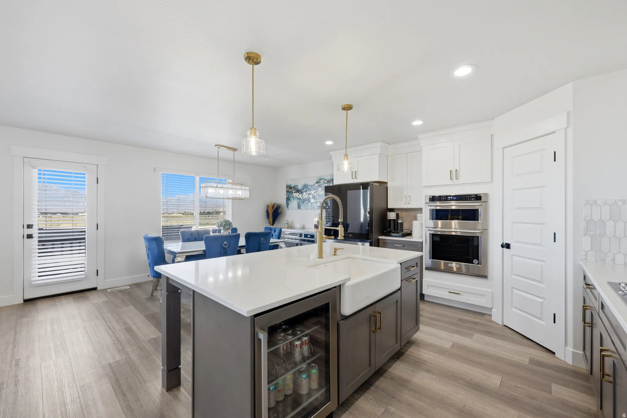 Kitchen featuring a large central island with a built-in beverage cooler and farmhouse sink, wood-finish flooring, white cabinetry, and stainless steel double ovens