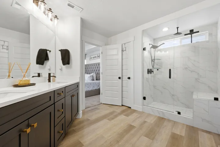 Spacious bathroom featuring wood-finish flooring, a dual vanity with dark cabinetry and white countertops, a large walk-in shower with glass enclosure and marble-look tile, and modern black fixtures