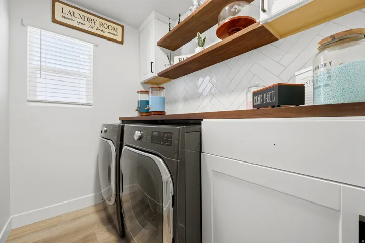 Dedicated laundry space featuring a window with blinds, wood-finish flooring, white cabinetry, open wood shelving, and a white herringbone tile backsplash