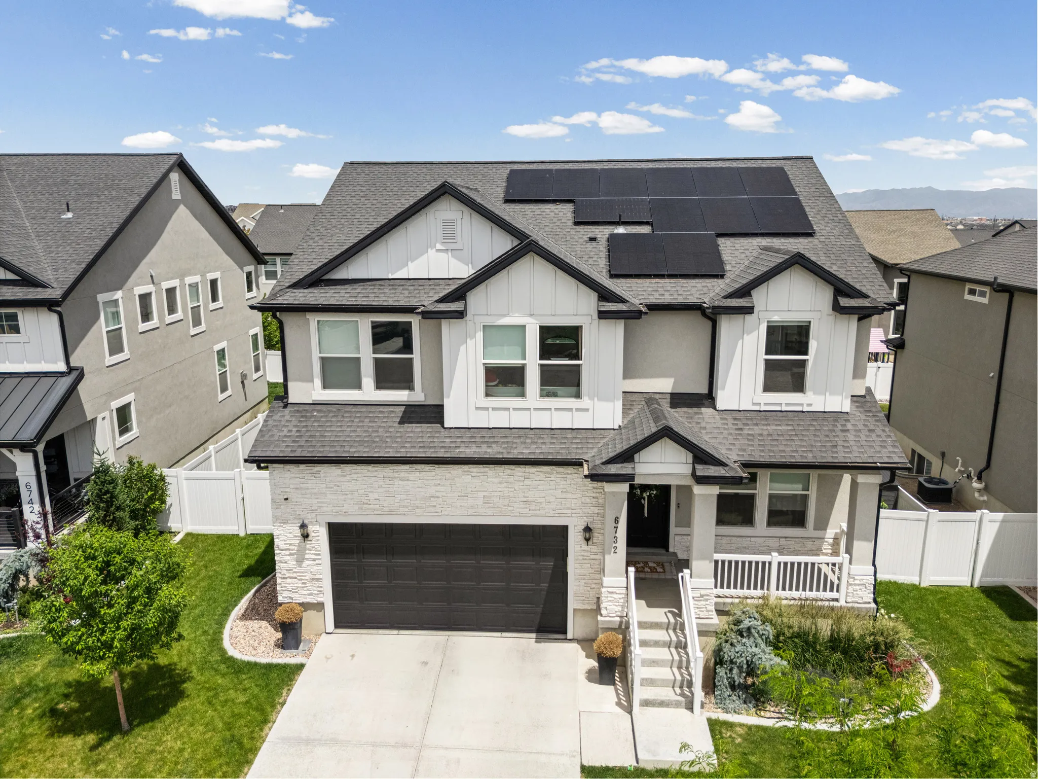 Modern residential exterior featuring a white board and batten siding, dark gray shingled roof with integrated solar panels, and a stone facade accenting the garage and front porch