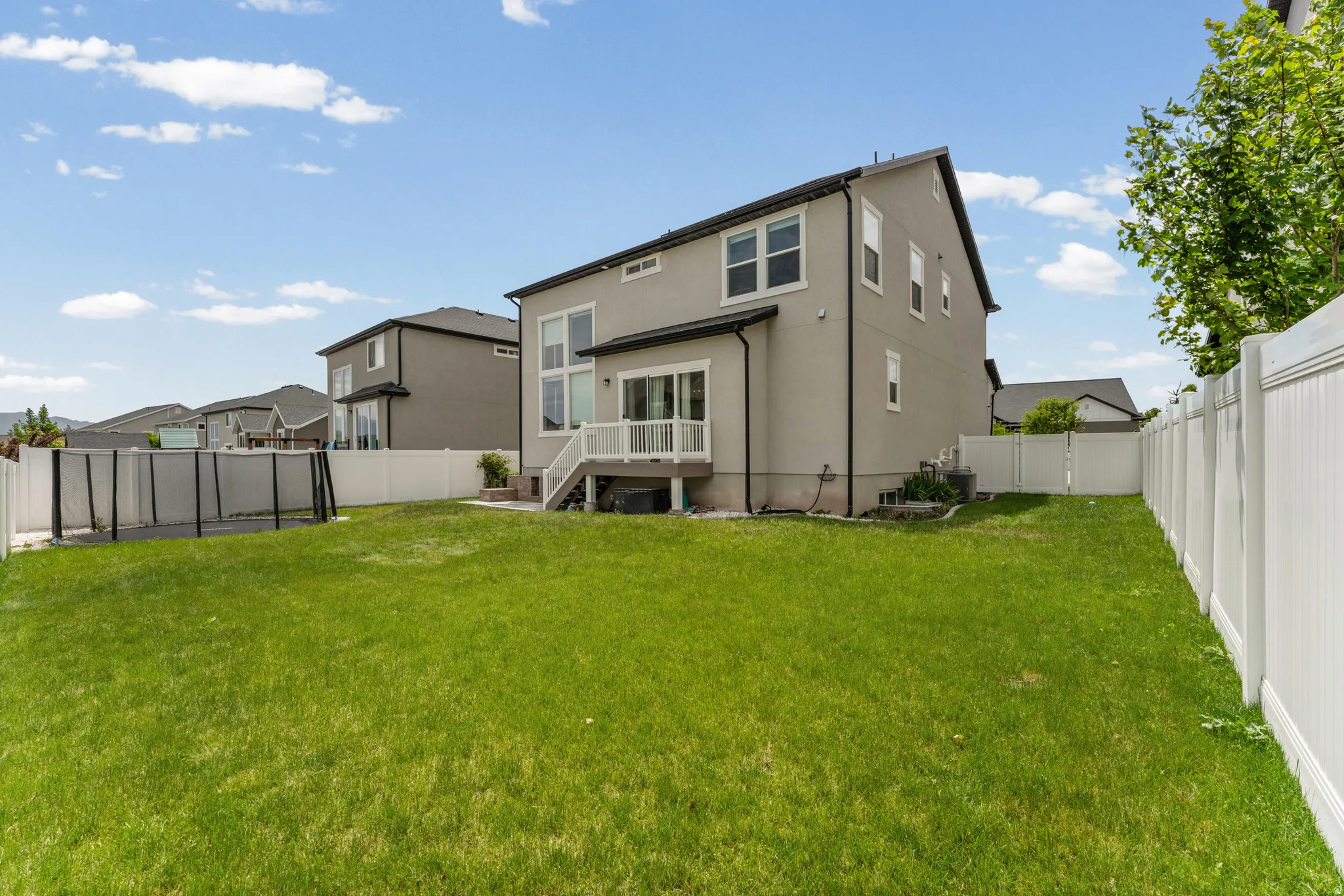 Expansive backyard featuring a lush green lawn, full privacy fencing, a ground-level deck, exterior stucco siding, and multiple windows