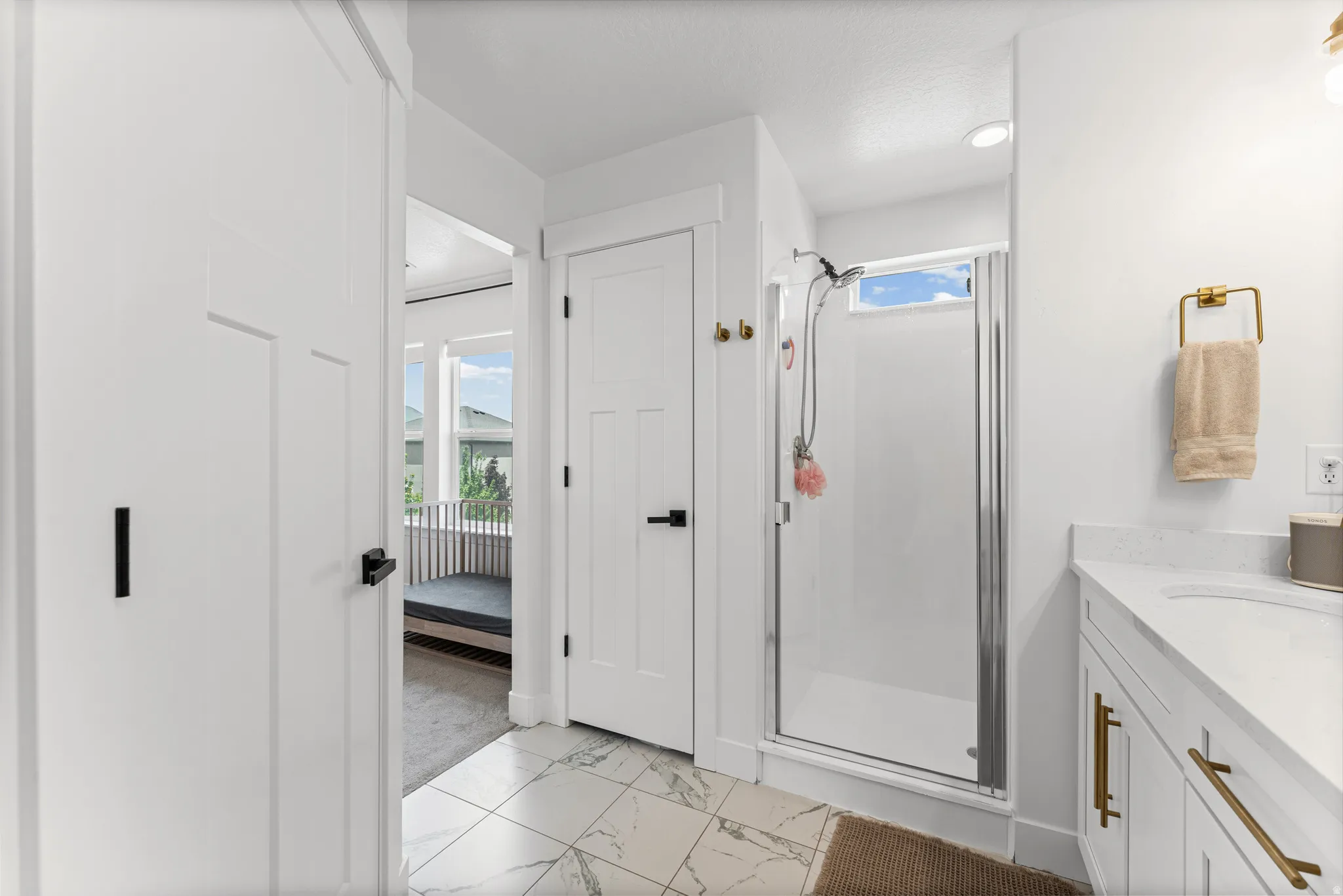 Bathroom featuring white paneled doors with black hardware, a shower with a clear glass enclosure and transom window, a single vanity with a white countertop and gold hardware, and white tile flooring