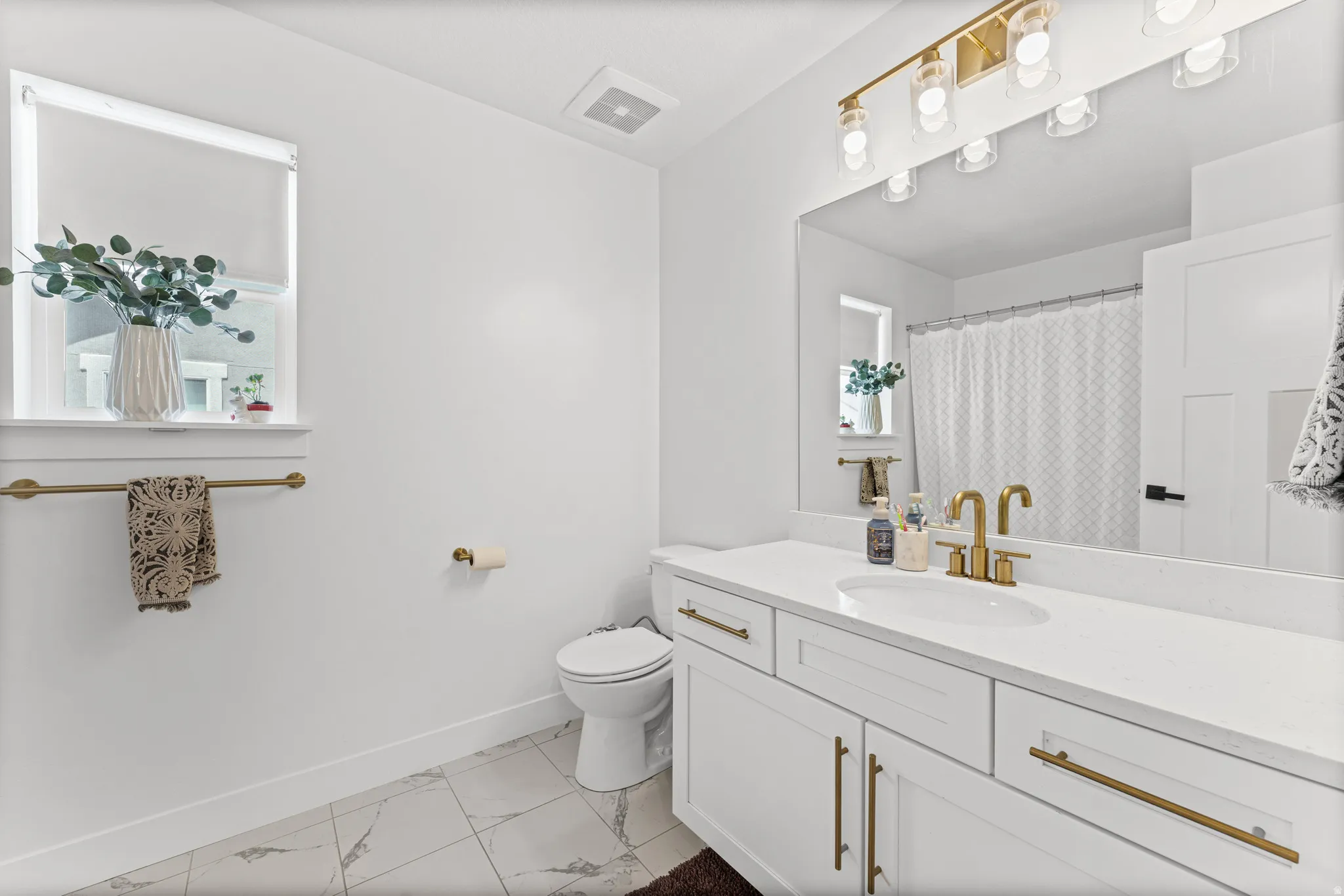 Modern bathroom featuring a white vanity with an integrated sink and brass-finish hardware, a large mirror, a multi-bulb vanity light with a brass-finish bar, white tiled flooring, and a white toilet