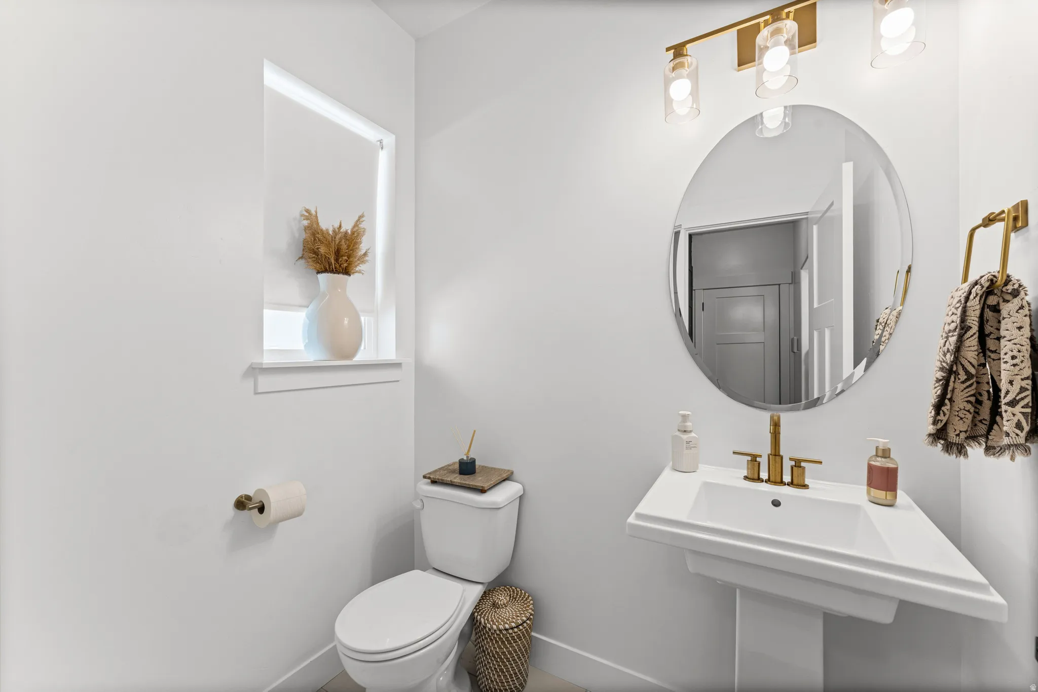 Modern powder room featuring a white pedestal sink with gold-tone fixtures, an oval mirror, and a three-light vanity fixture