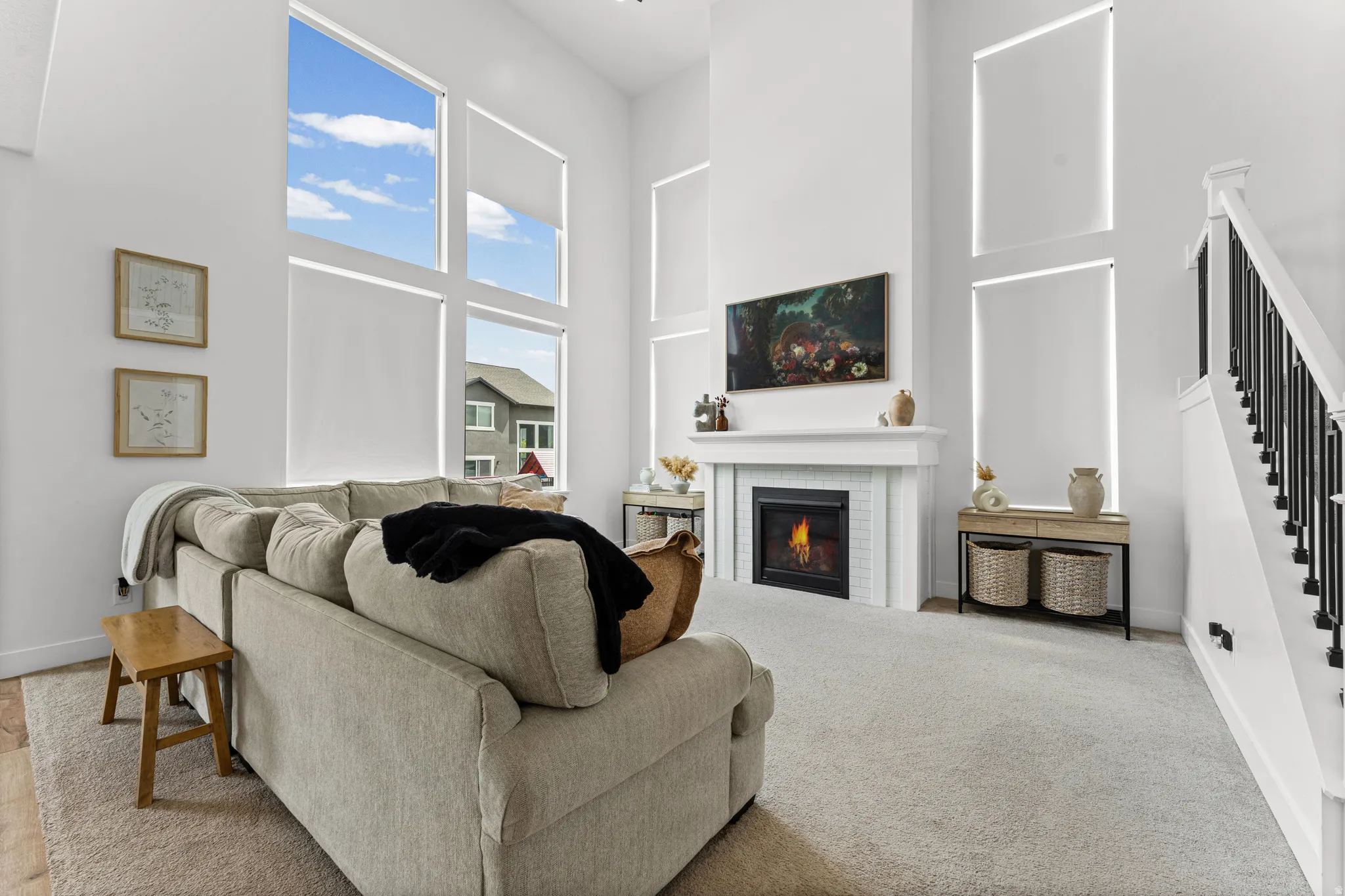 Two-story living area featuring a tile-surround fireplace with a classic mantel, a two-story window wall with roller shades, high ceilings, a carpeted floor, and a staircase with black metal balusters