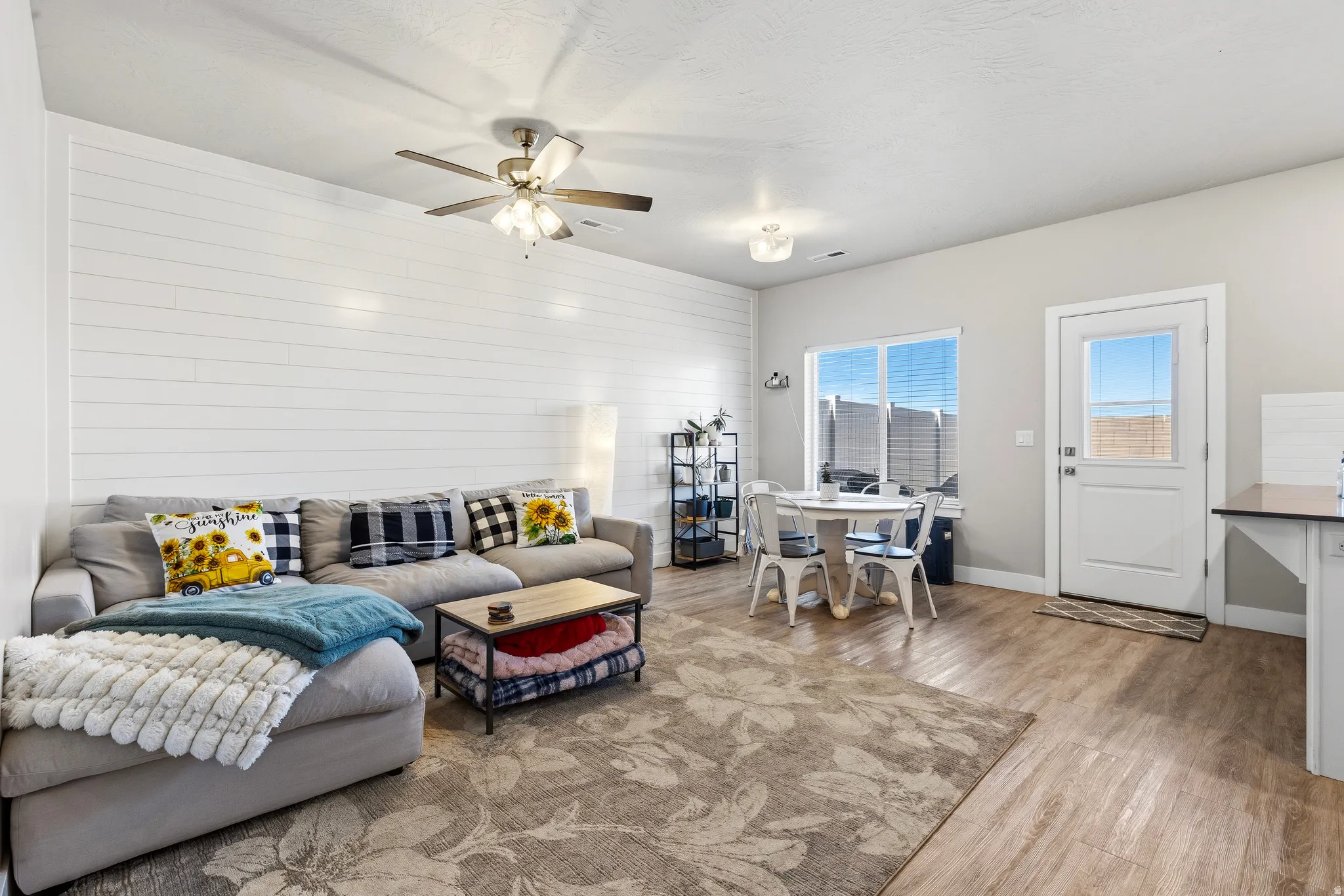 Open-concept living space featuring a shiplap accent wall, wood-finish flooring, a ceiling fan with integrated lighting, and a dining area with direct outdoor access
