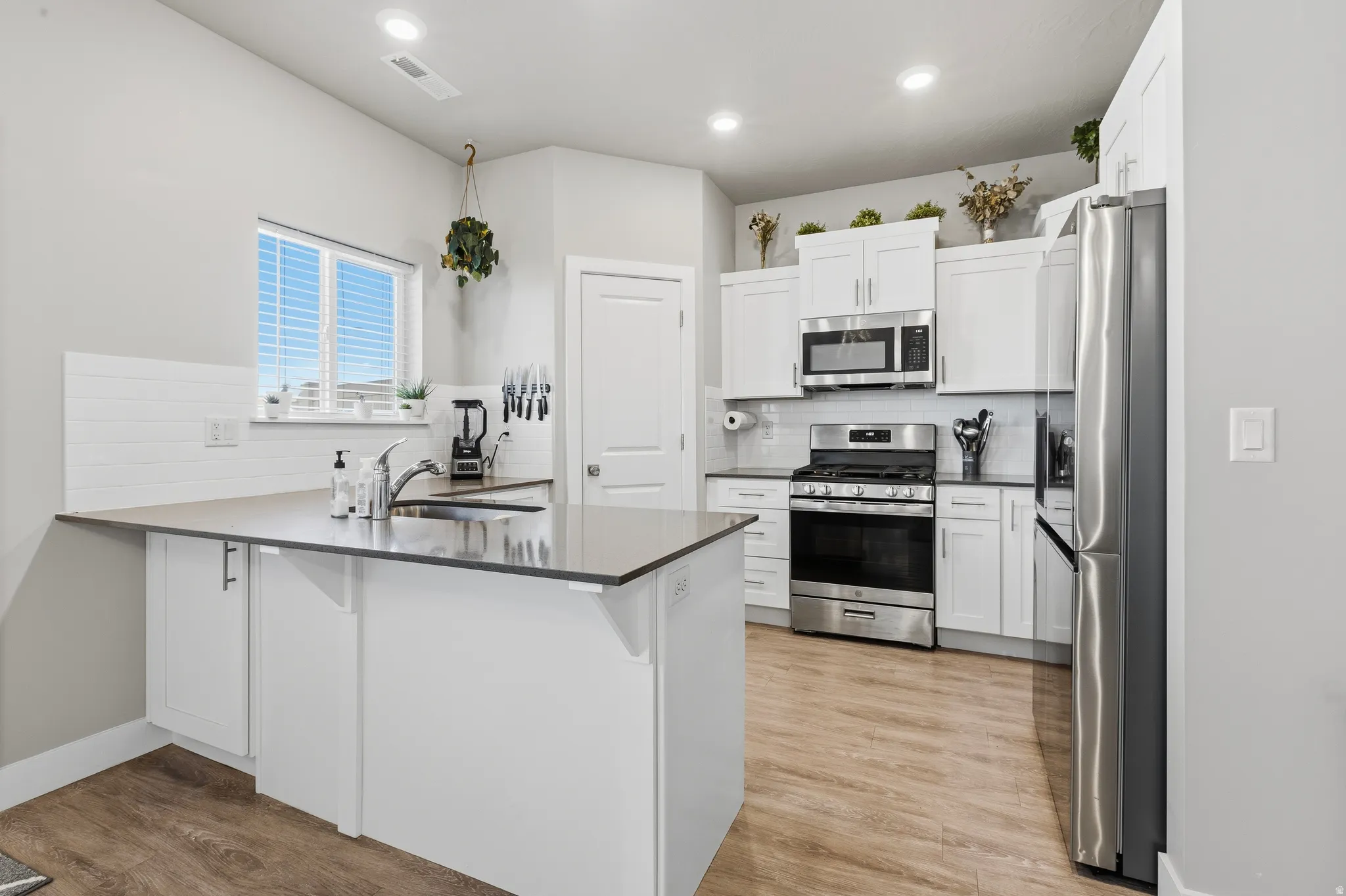 Kitchen featuring an island with an undermount sink, white cabinetry, stainless steel appliances, wood-finish flooring, and a white subway tile backsplash