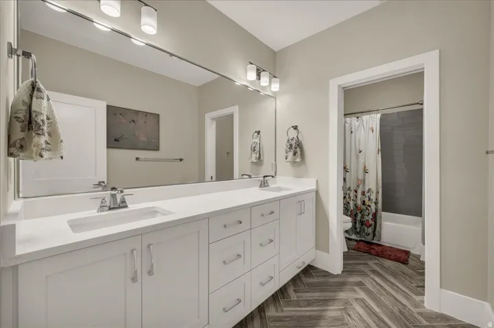 Bathroom featuring a spacious double vanity with white cabinetry and integrated sinks, a large wall-mounted mirror, overhead bar lighting, and wood-finish flooring laid in a herringbone pattern