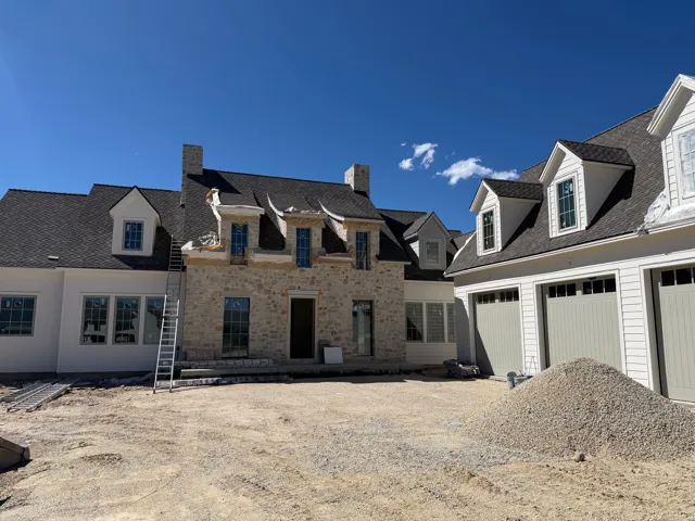 Stone facade, multiple dormer windows, dark shingle roof, multi-pane windows, light-toned garage doors