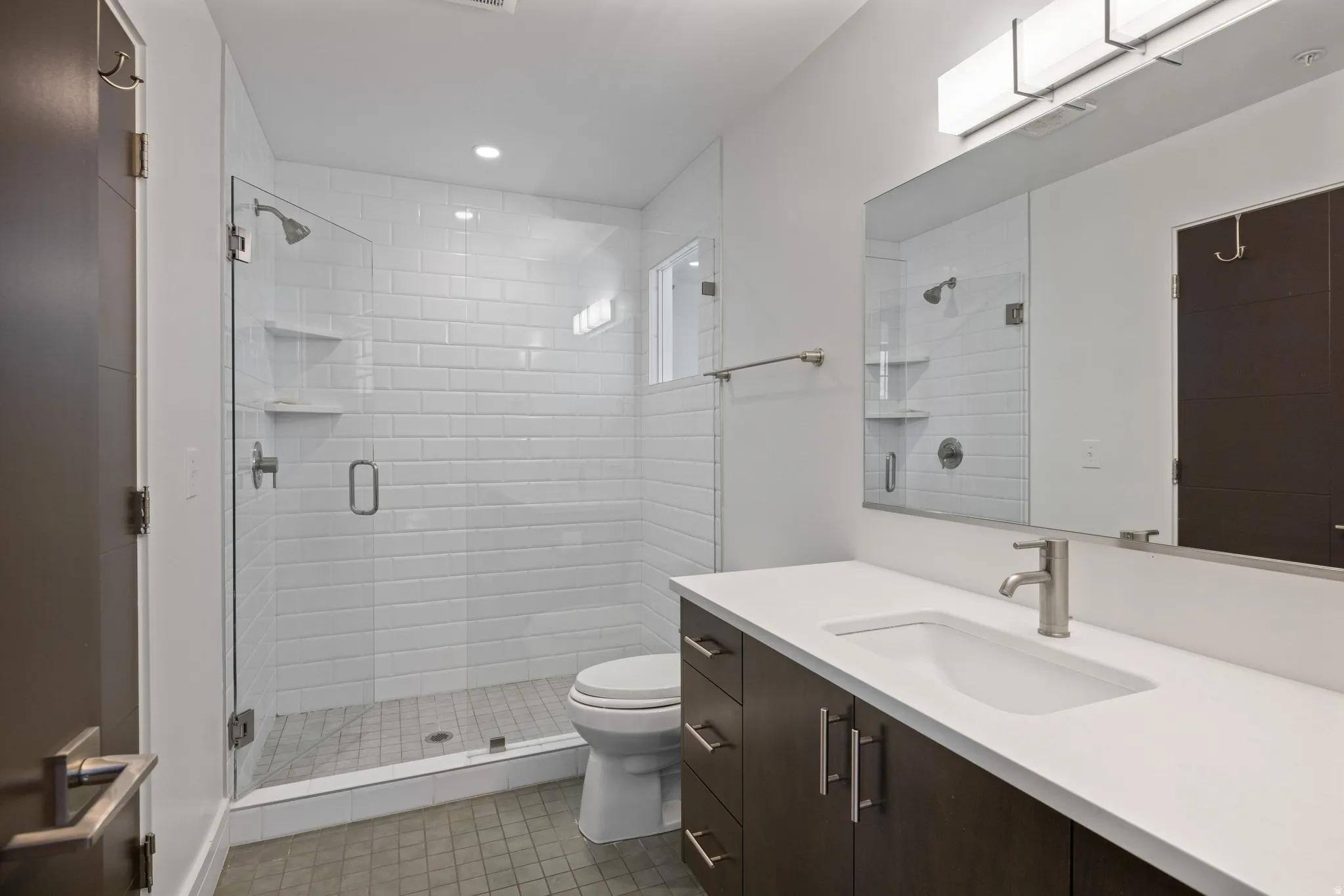 Contemporary bathroom featuring a dark wood-finish vanity with a white countertop and integrated sink, a frameless mirror, and a glass-enclosed shower with white subway tile