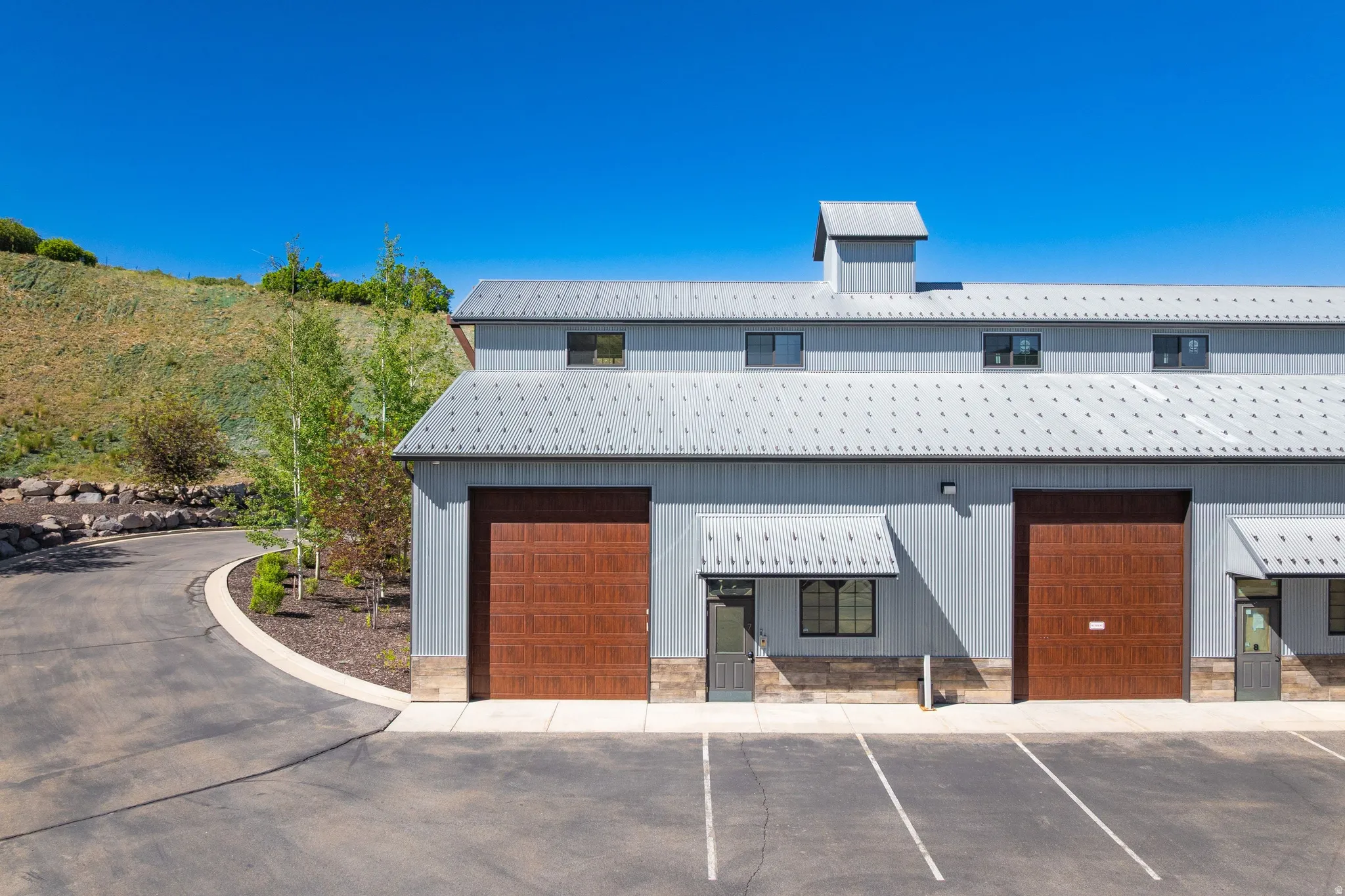 Modern industrial-style structure featuring a light gray exterior, two expansive brown garage doors, multiple windows with awnings, a cupola, and a paved parking area