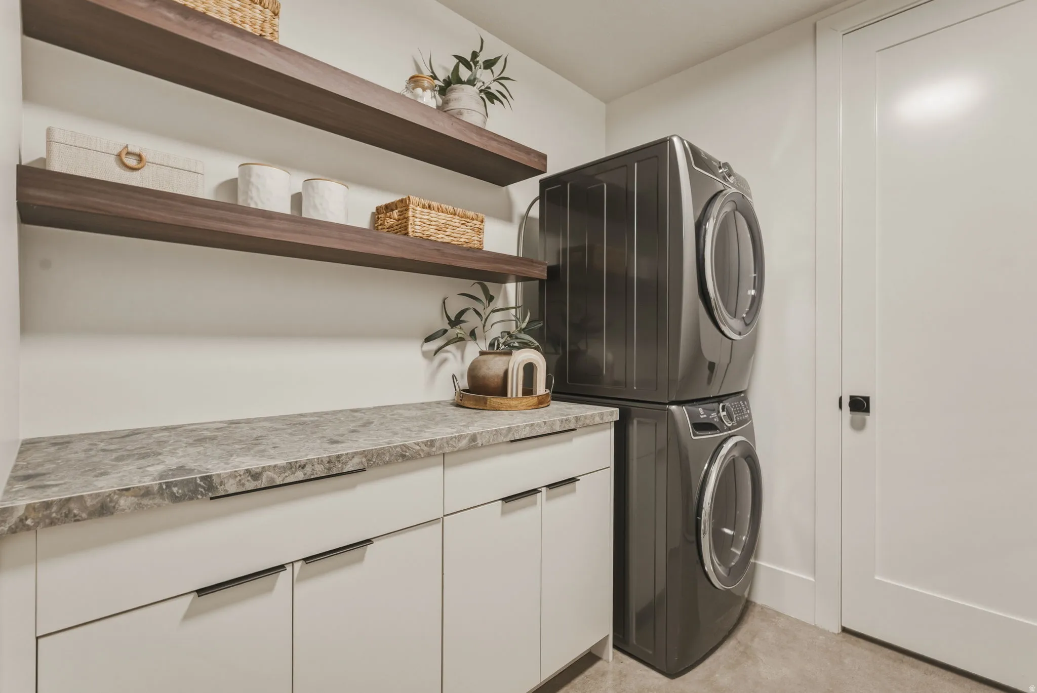 Modern laundry area featuring a stacked washer and dryer, white cabinetry, a patterned countertop, and dual wood-finish floating shelves
