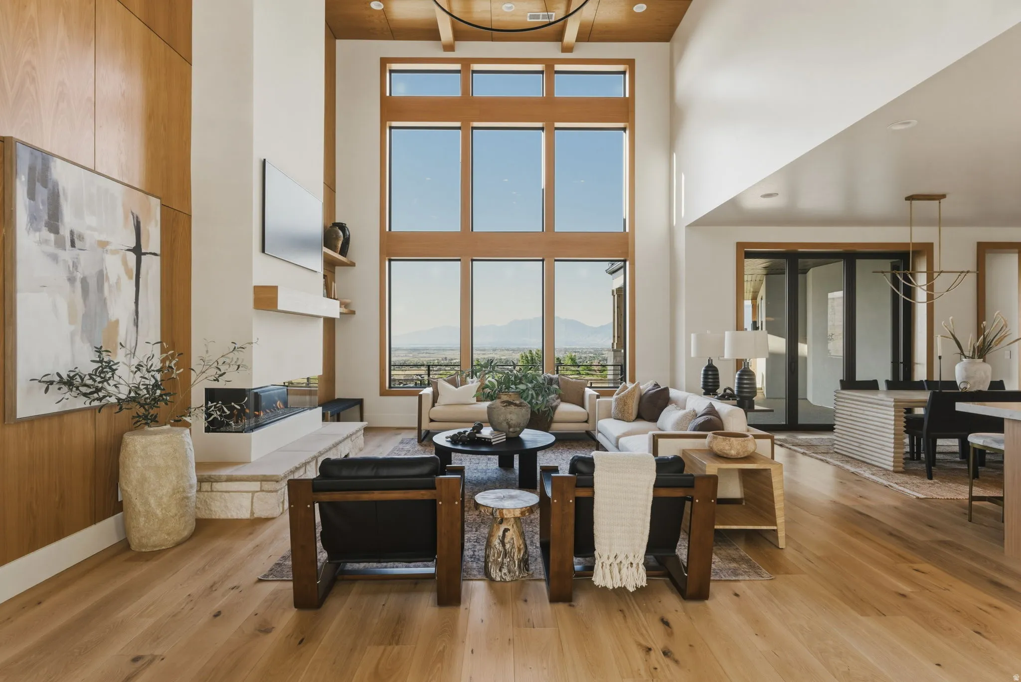 Two-story living area featuring extensive window walls, wood-finish flooring, a linear fireplace with stone hearth, a wood-paneled accent wall, and a wood-slat ceiling with exposed beams