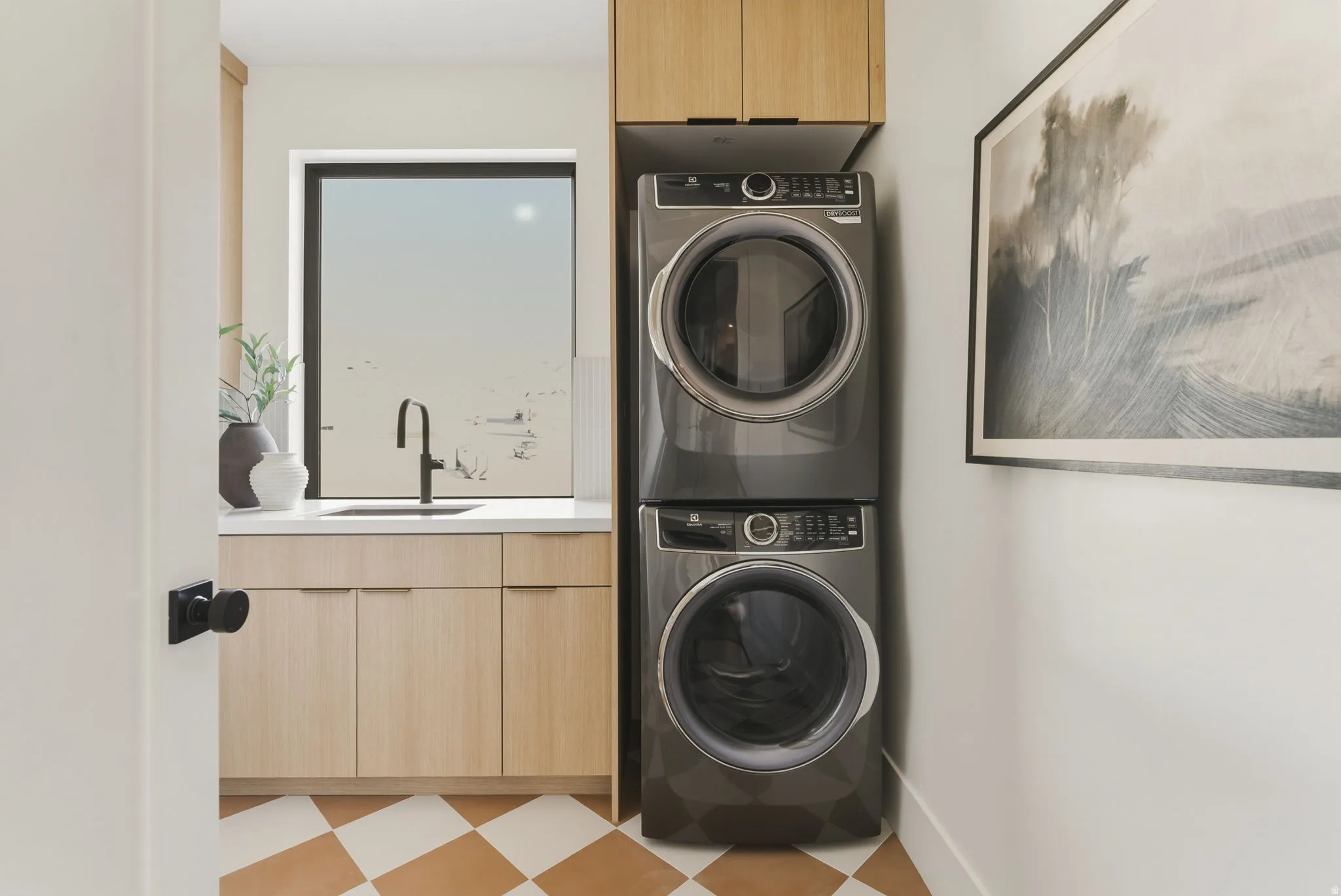 Laundry room featuring a stacked washer and dryer, light wood-finish cabinetry, a white countertop with an undermount sink, a black gooseneck faucet, and checkerboard floor tiling