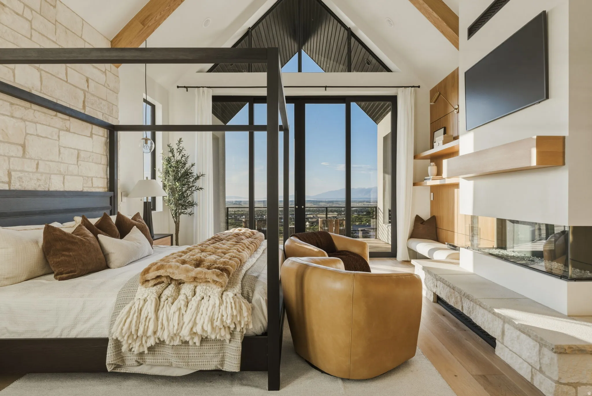 Vaulted ceiling with exposed wood beams, stone accent wall, floor-to-ceiling windows, built-in fireplace, and light wood-finish flooring