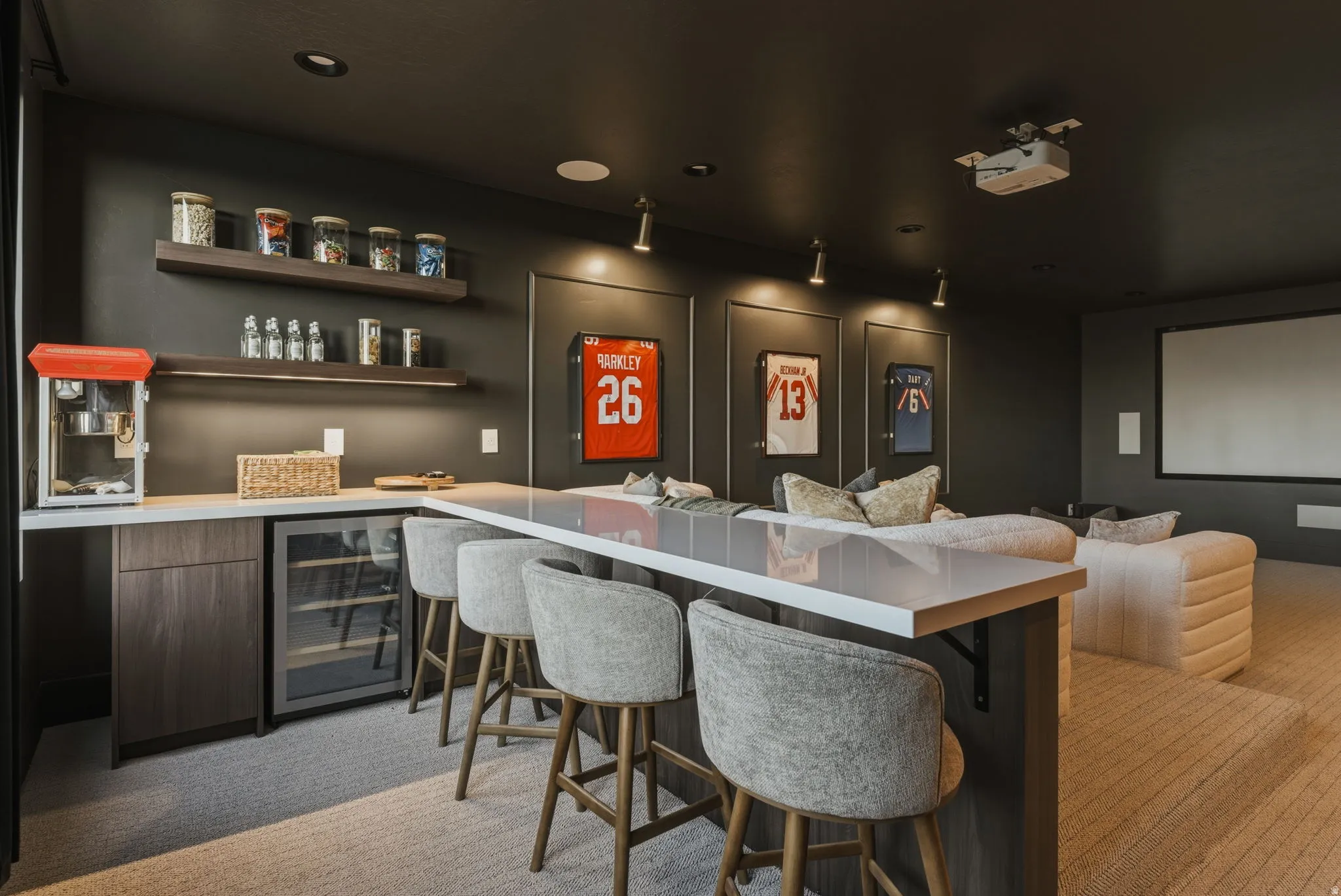 Dedicated entertainment room featuring a built-in bar with a beverage refrigerator, layered floating shelves with integrated lighting, and a large projection screen