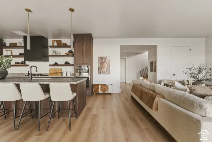 Open-concept living area featuring wood-finish flooring, a kitchen island with seating, dark wood cabinetry, integrated oven, and a modern range hood