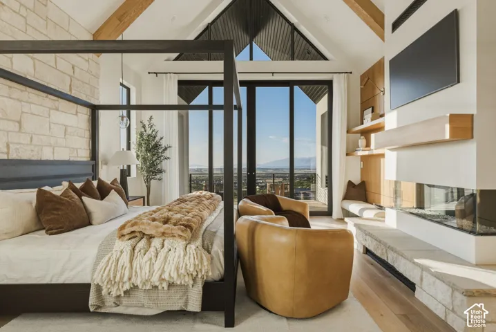 Vaulted ceiling with exposed wood beams, stone accent wall, floor-to-ceiling windows, built-in fireplace, and light wood-finish flooring
