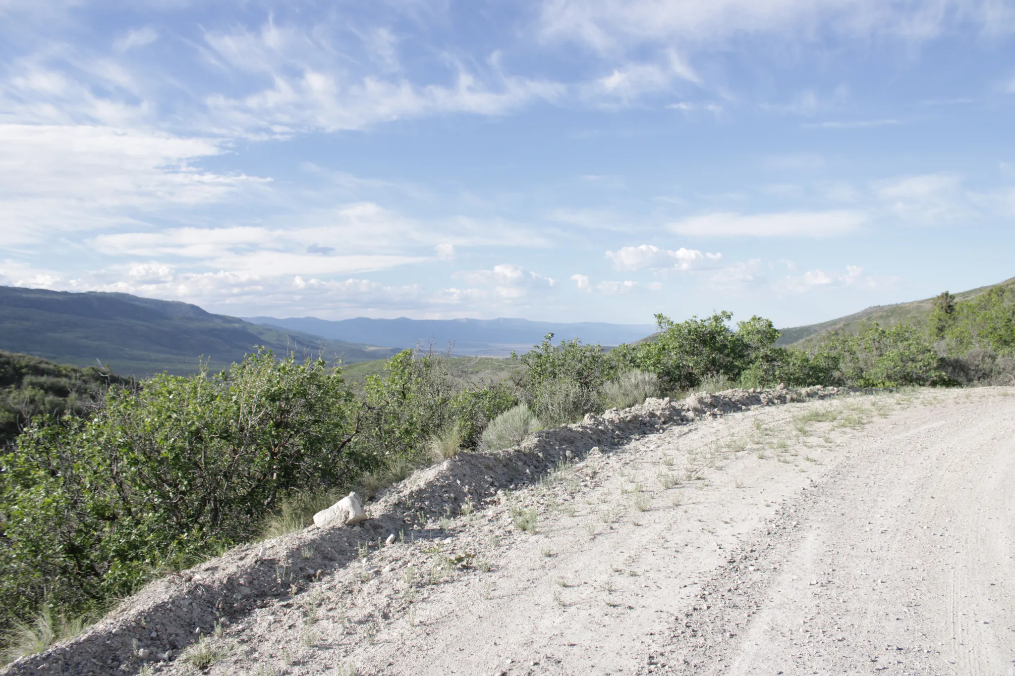 Expansive mountain landscape with a partial dirt road, natural foliage, and distant valleys under a blue sky