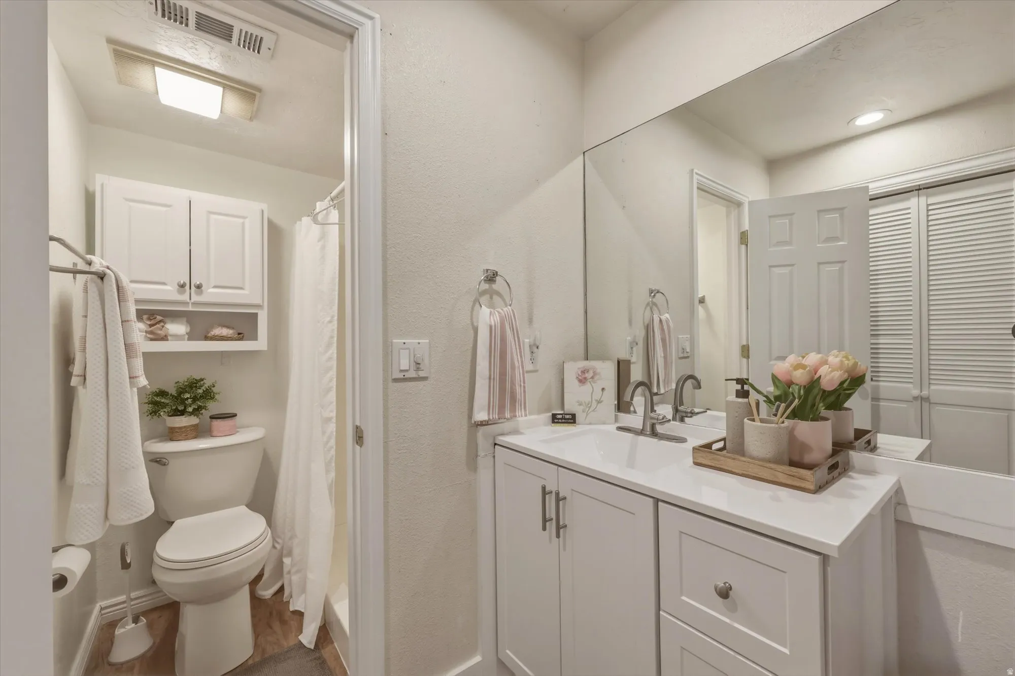 Bathroom featuring a white vanity with a single sink, an expansive wall-mounted mirror, white shaker-style cabinetry with silver hardware, a toilet, and a shower enclosure