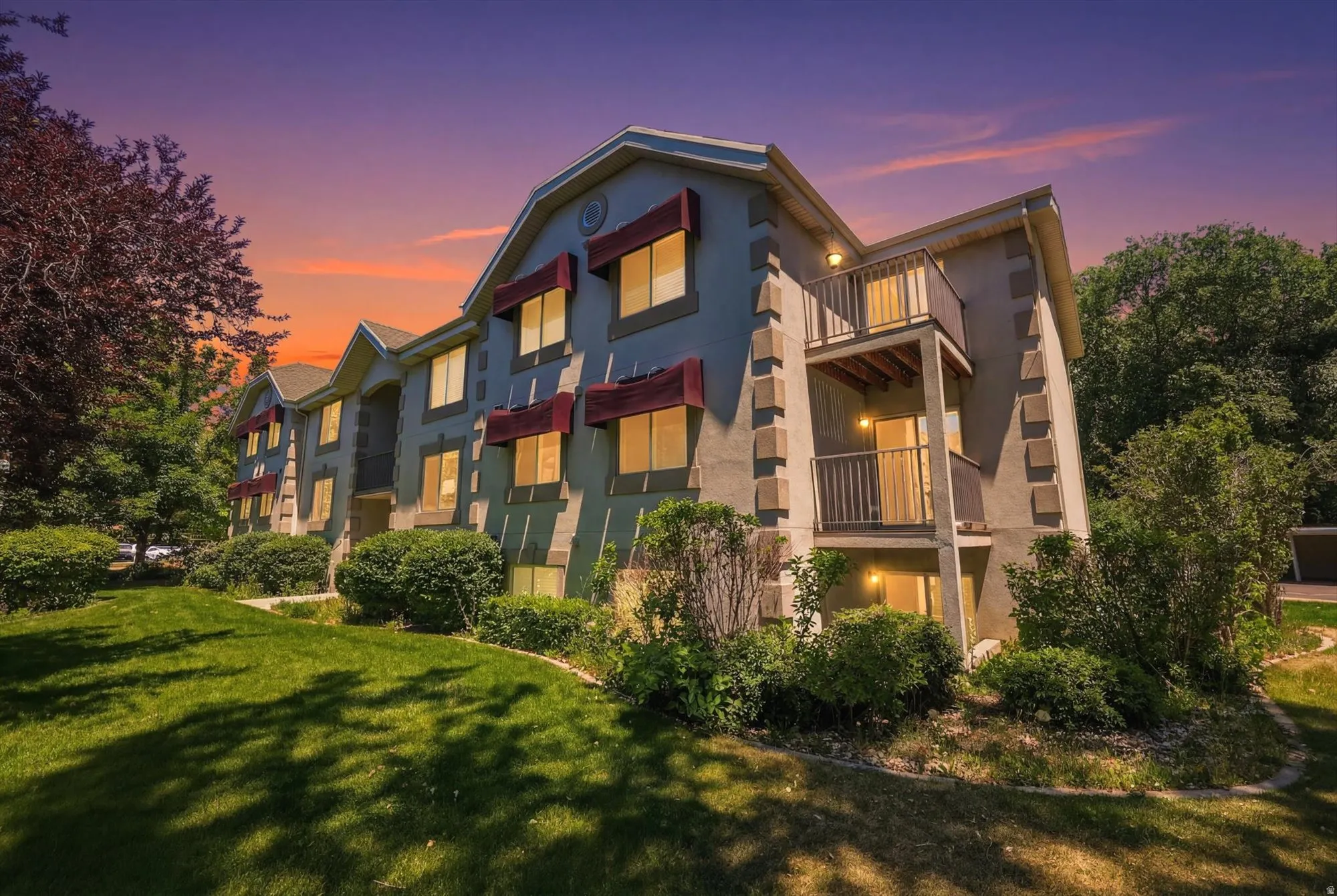 Multi-story building featuring stucco exterior, multiple windows with red awnings, decorative stone-look corner accents, and private balconies with metal railings