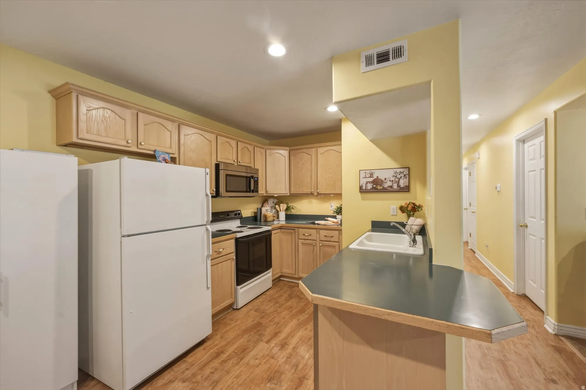 Kitchen featuring wood-finish flooring, light wood cabinetry, white appliances, a built-in microwave, and a breakfast bar with a sink