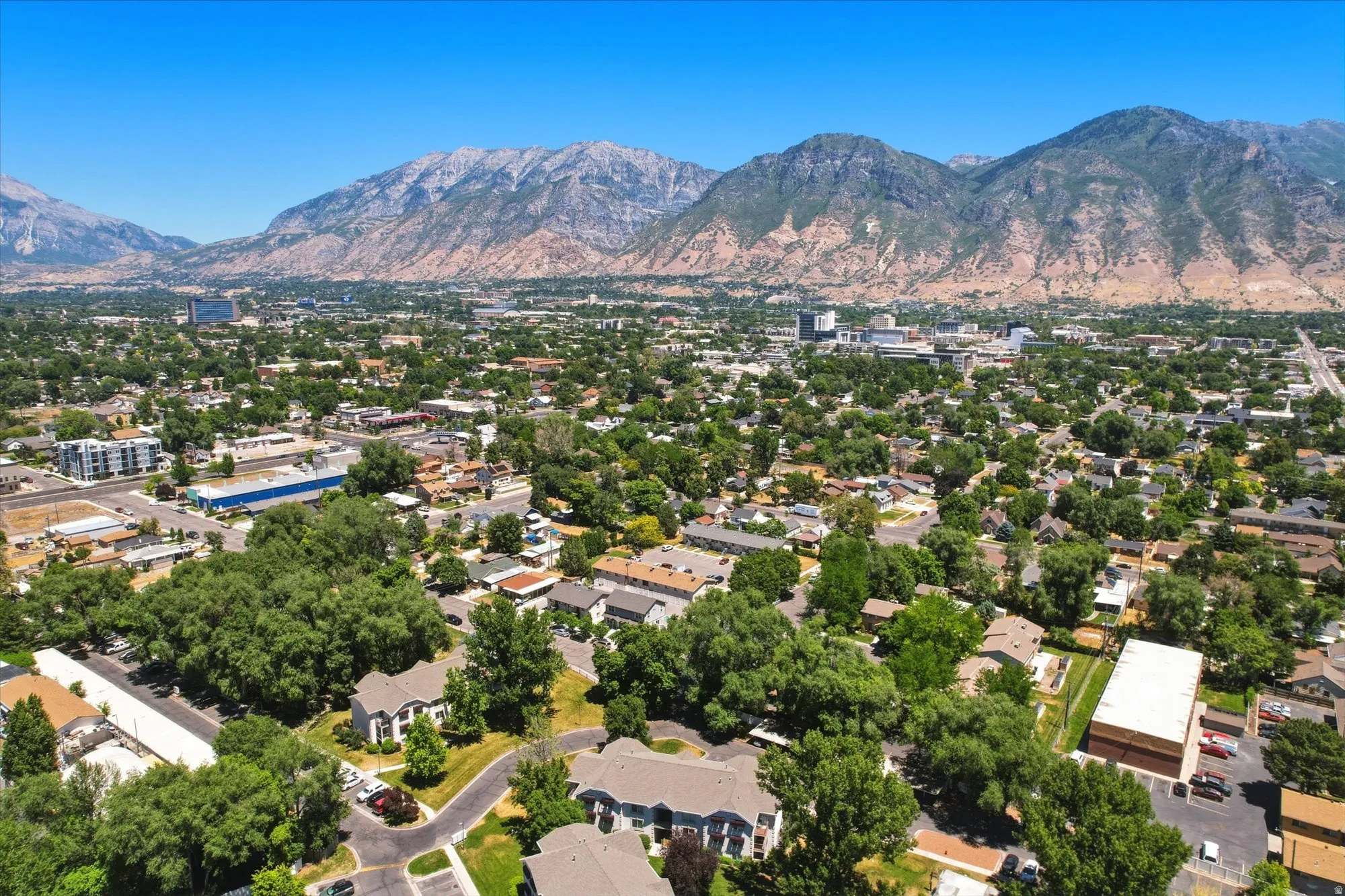 Expansive aerial perspective showcasing a mountainous backdrop, a clear blue sky, abundant mature trees, a mix of residential and commercial structures, and defined roadways