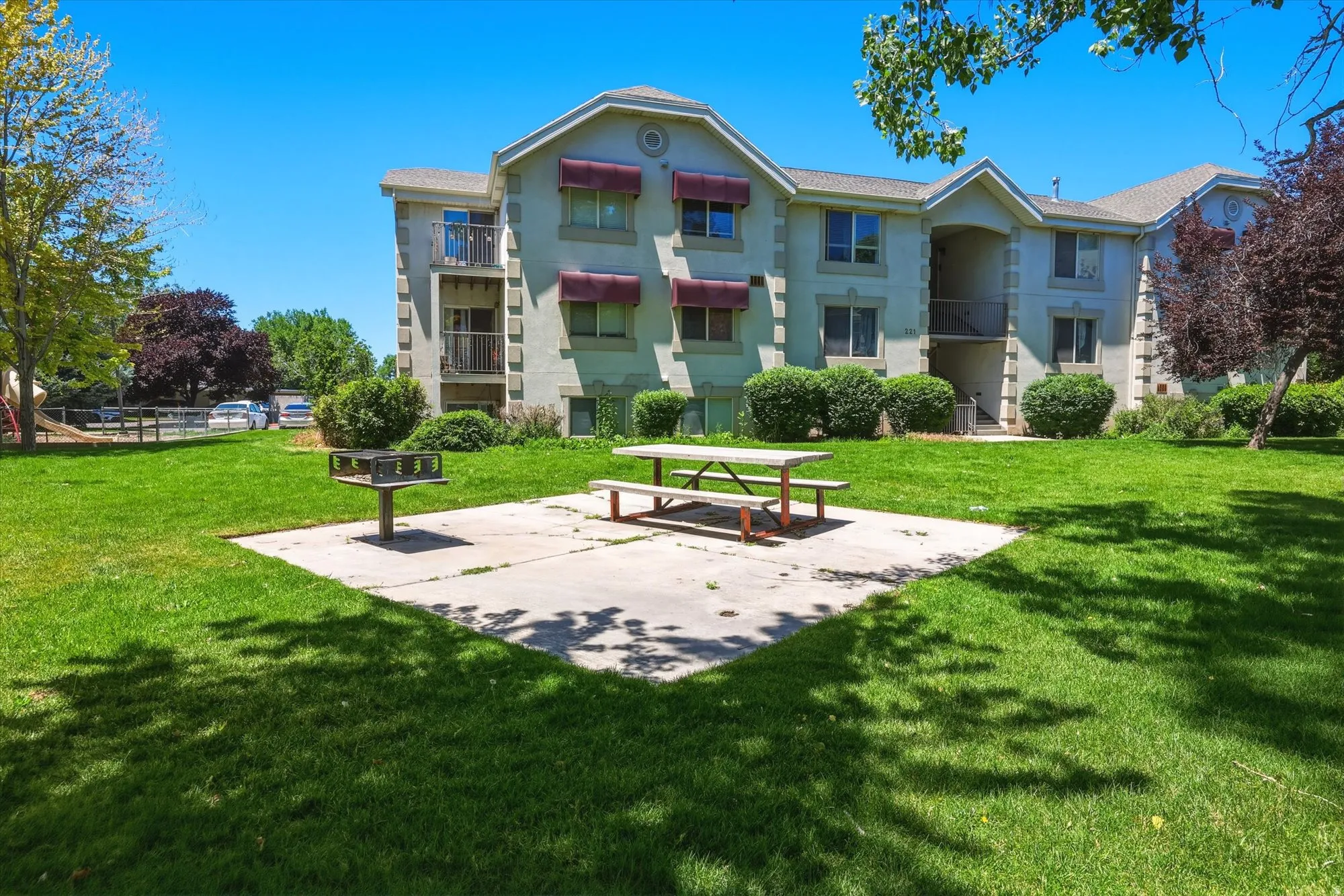 Spacious green lawn with a concrete patio featuring a picnic table and a built-in barbecue grill, bordered by mature landscaping and a multi-story building with balconies and red awnings