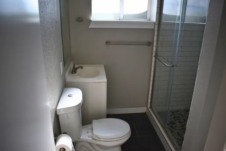 Bathroom featuring a white vanity with integrated sink, toilet, glass-enclosed shower with subway tile surround, and dark tile flooring