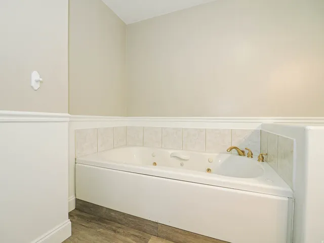Corner soaking tub with brass fixtures and tiled surround, featuring wainscoting and wood-finish flooring