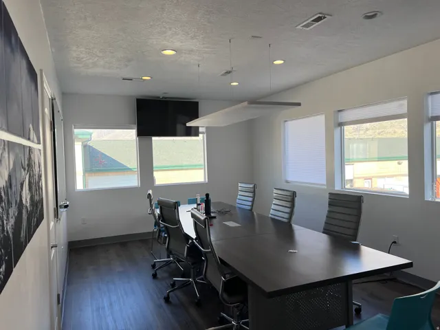 Spacious meeting room featuring wood-finish flooring, multiple windows with blinds, recessed ceiling lights, and a wall-mounted display