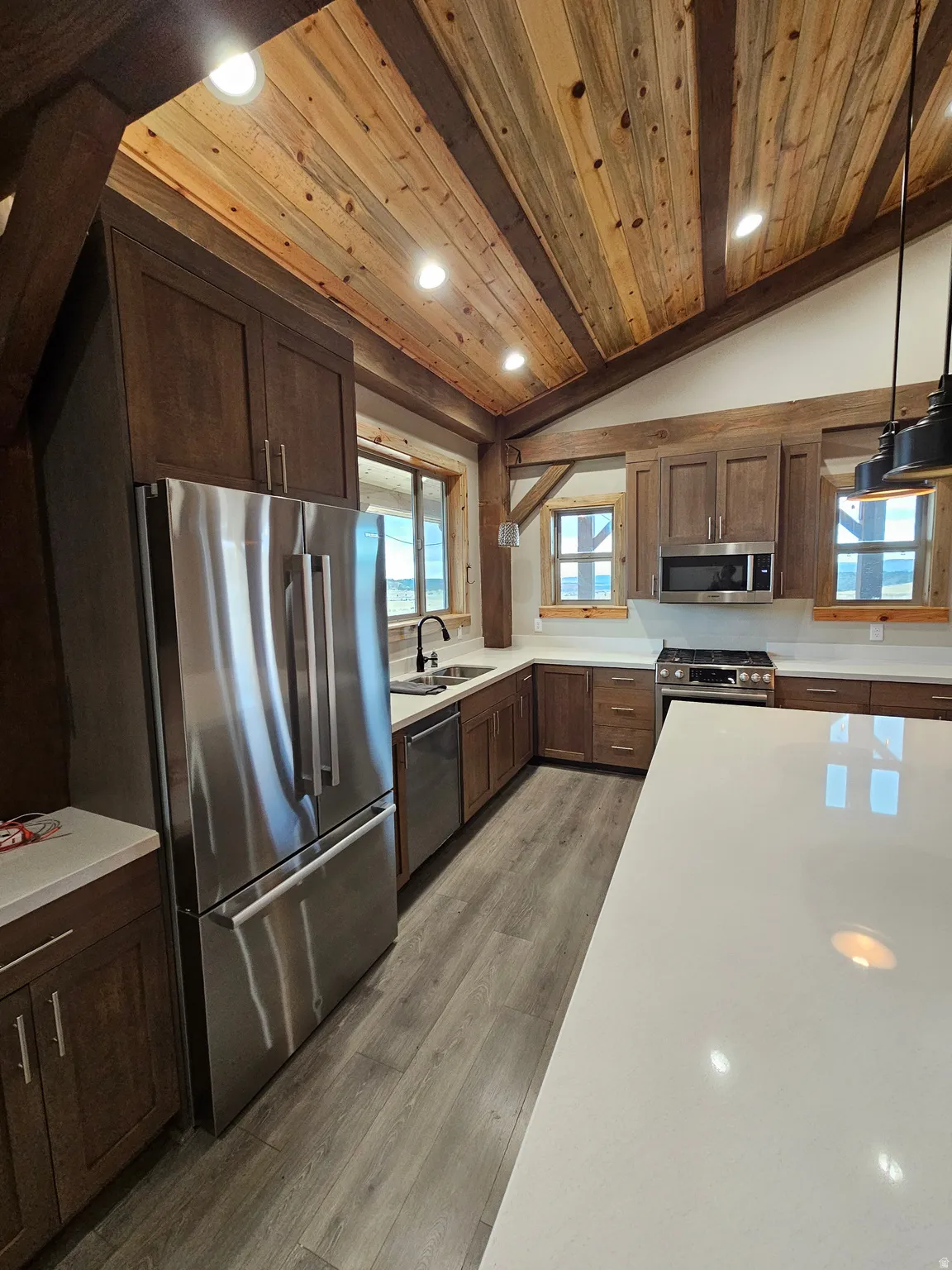 Kitchen featuring a vaulted wood ceiling with exposed wood beams, a central island with a light-toned countertop, dark wood-finish cabinetry, stainless steel appliances, and wood-finish flooring