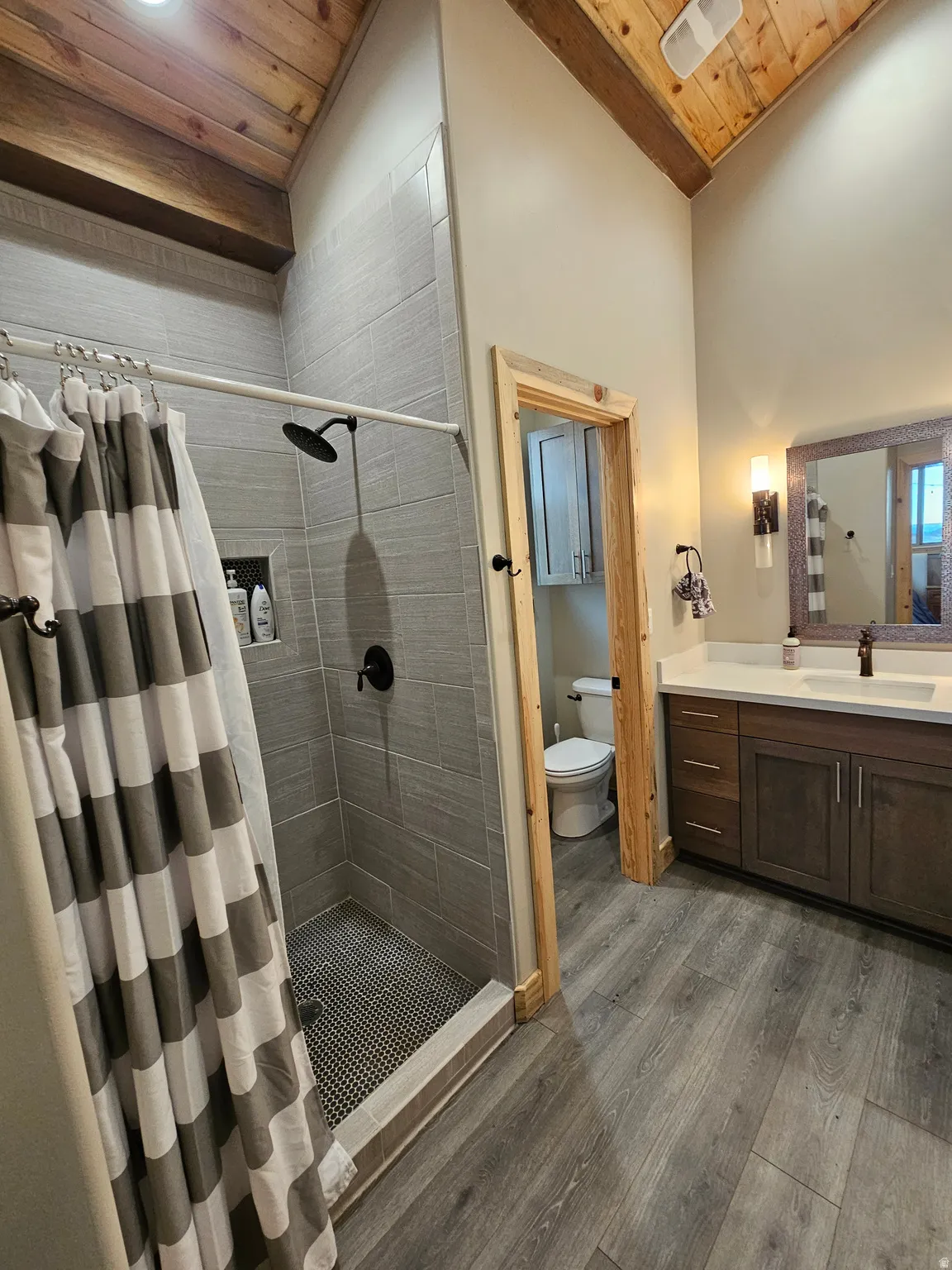 Spacious bathroom featuring a tiled shower with matte black fixtures, dual vanity with a light countertop, wood-finish flooring, and a vaulted wood-paneled ceiling with exposed beams