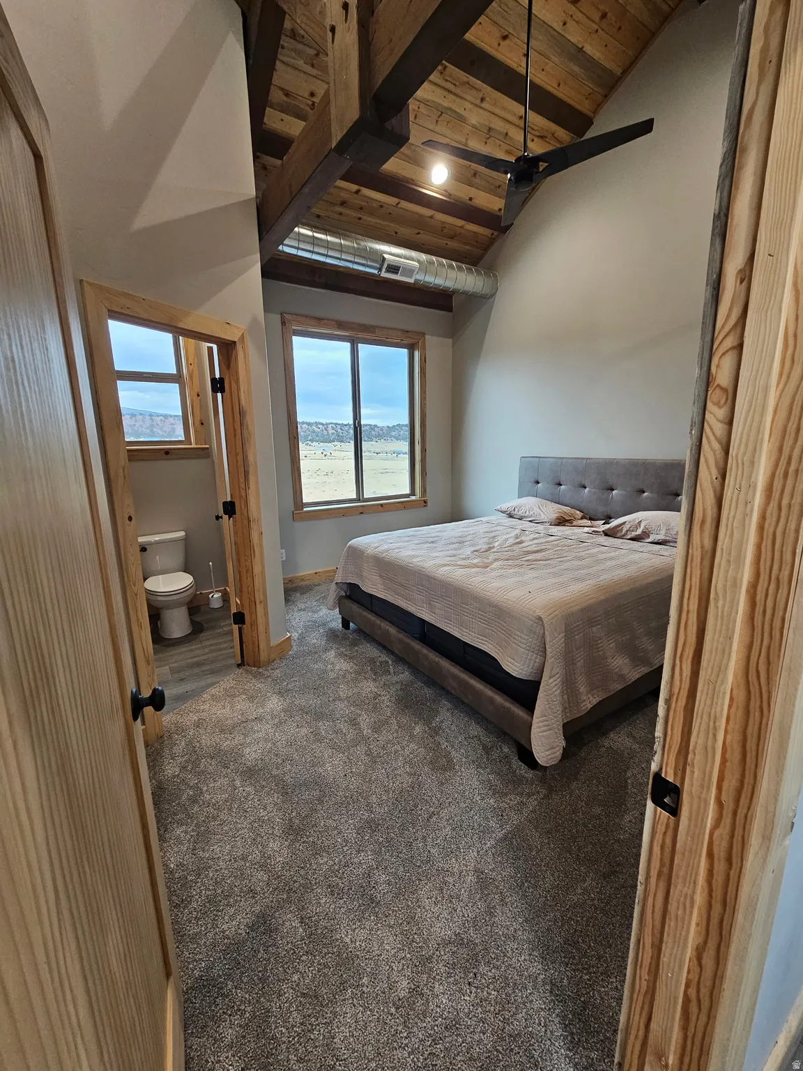 Bedroom featuring a vaulted wood ceiling with exposed beams, wood-trimmed windows, a ceiling fan, and attached bathroom