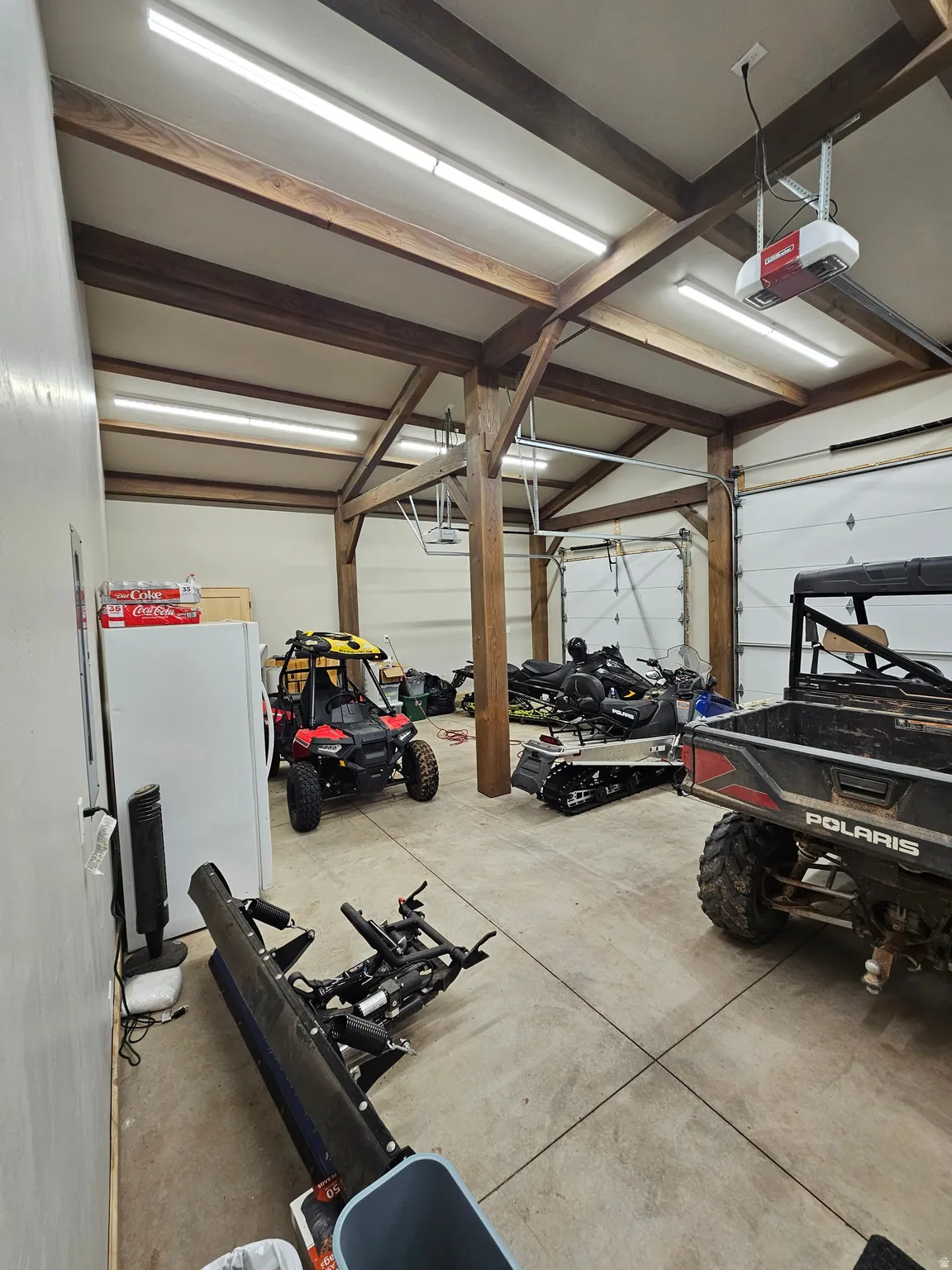Spacious garage featuring exposed wood beams, multiple overhead fluorescent light fixtures, two overhead garage doors with automatic openers, and a concrete floor