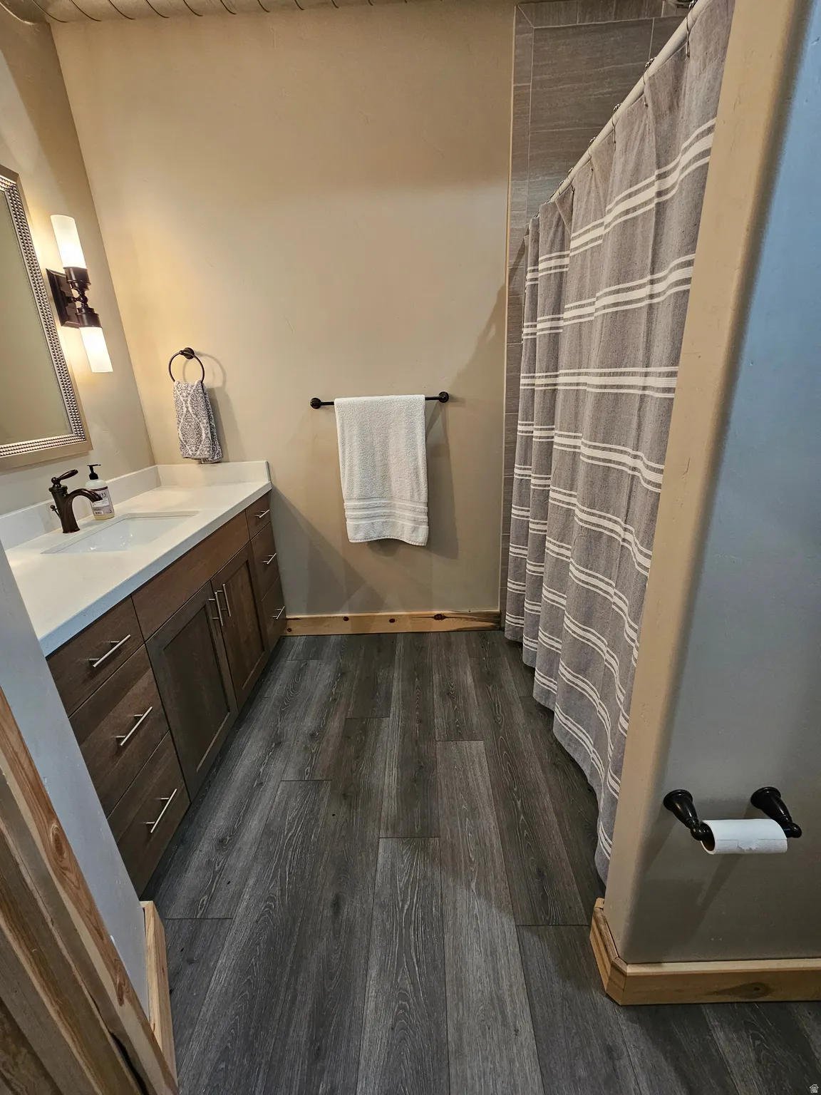 Bathroom featuring wood-finish flooring, a contemporary vanity with a white countertop and rectangular sink, a wall-mounted light fixture, and a tiled shower surround