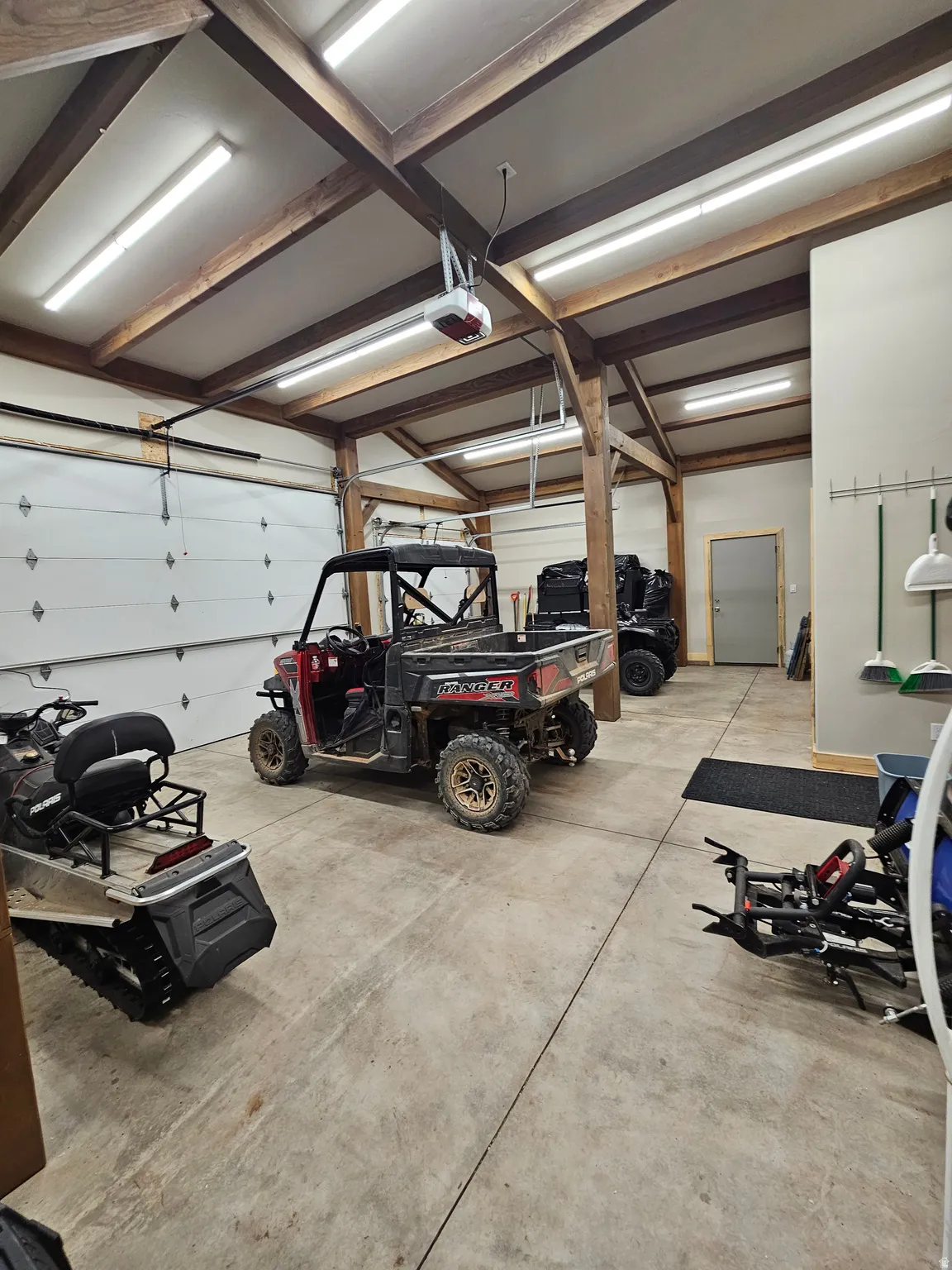 Spacious garage featuring exposed wood beams, concrete flooring, multiple overhead garage doors, abundant overhead lighting, and a service door