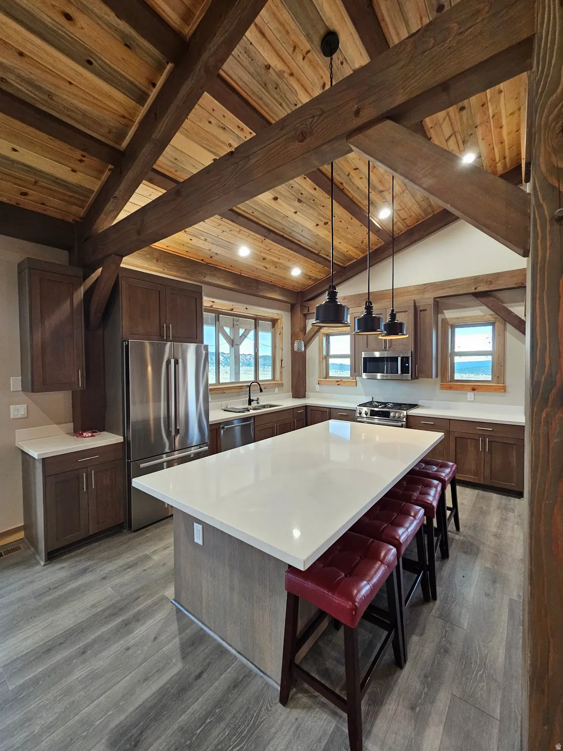 Kitchen featuring a vaulted wood ceiling with exposed wood beams, a central island with a light-toned countertop, dark wood-finish cabinetry, stainless steel appliances, and wood-finish flooring