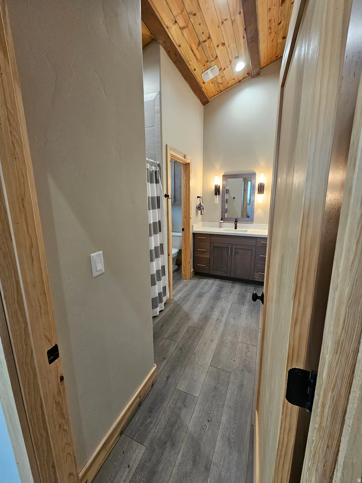 Spacious bathroom featuring a tiled shower with matte black fixtures, dual vanity with a light countertop, wood-finish flooring, and a vaulted wood-paneled ceiling with exposed beams