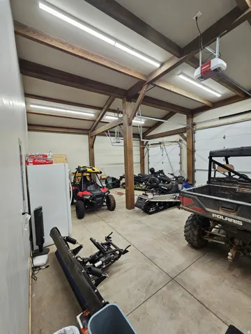 Spacious garage featuring exposed wood beams, multiple overhead fluorescent light fixtures, two overhead garage doors with automatic openers, and a concrete floor